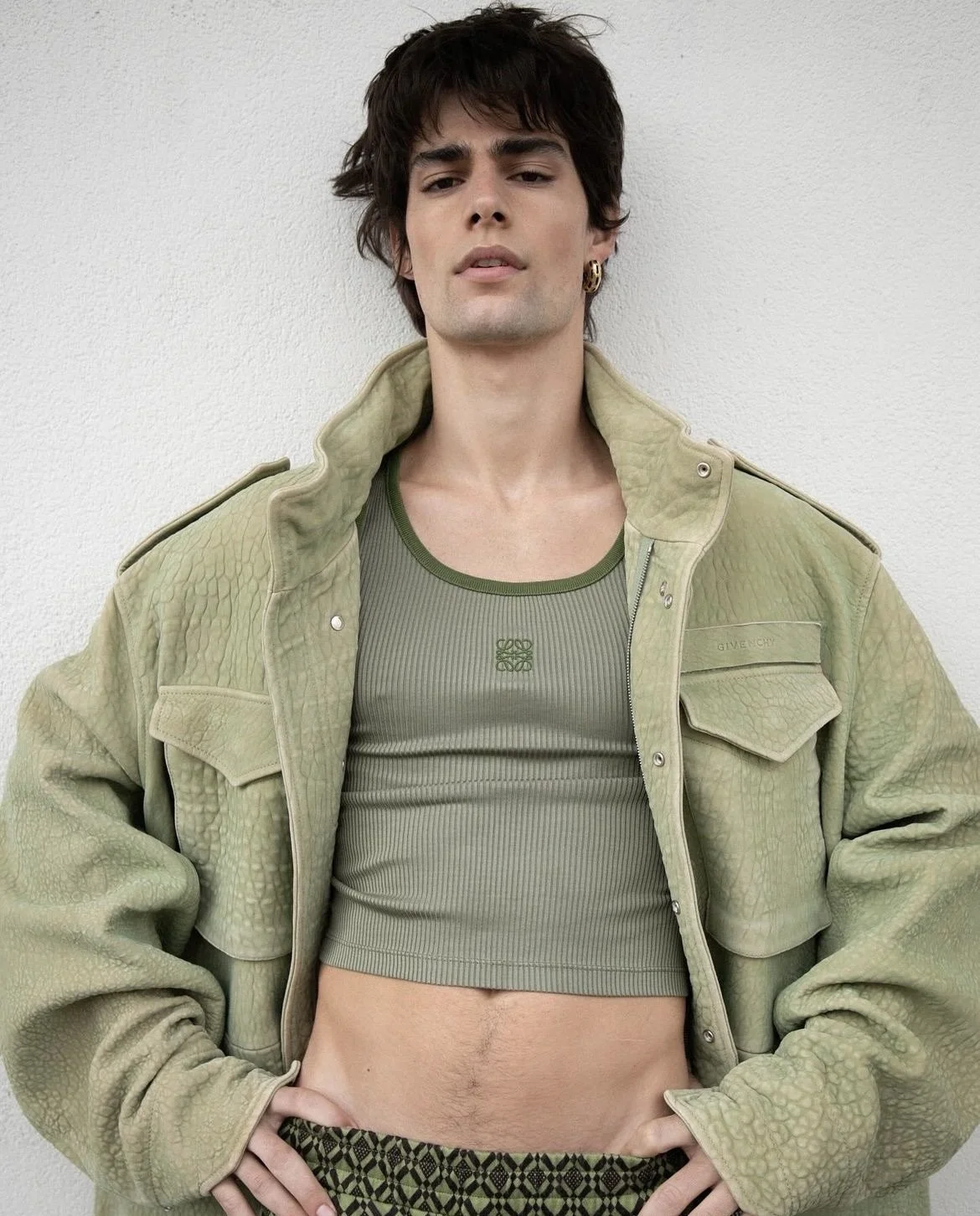 A young person with dark, tousled hair, wearing a green textured jacket open over a matching green crop top, and patterned pants, standing against a plain white wall.