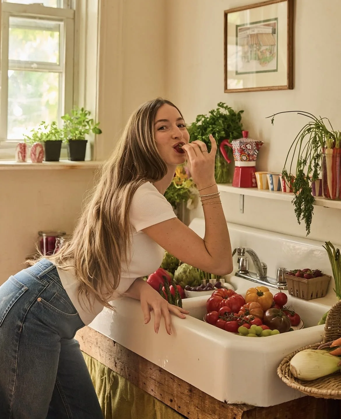 A woman with long brown hair wearing a white shirt and jeans standing at a kitchen sink filled with fresh tomatoes and vegetables, eating a snack, with a window and potted plants in the background.