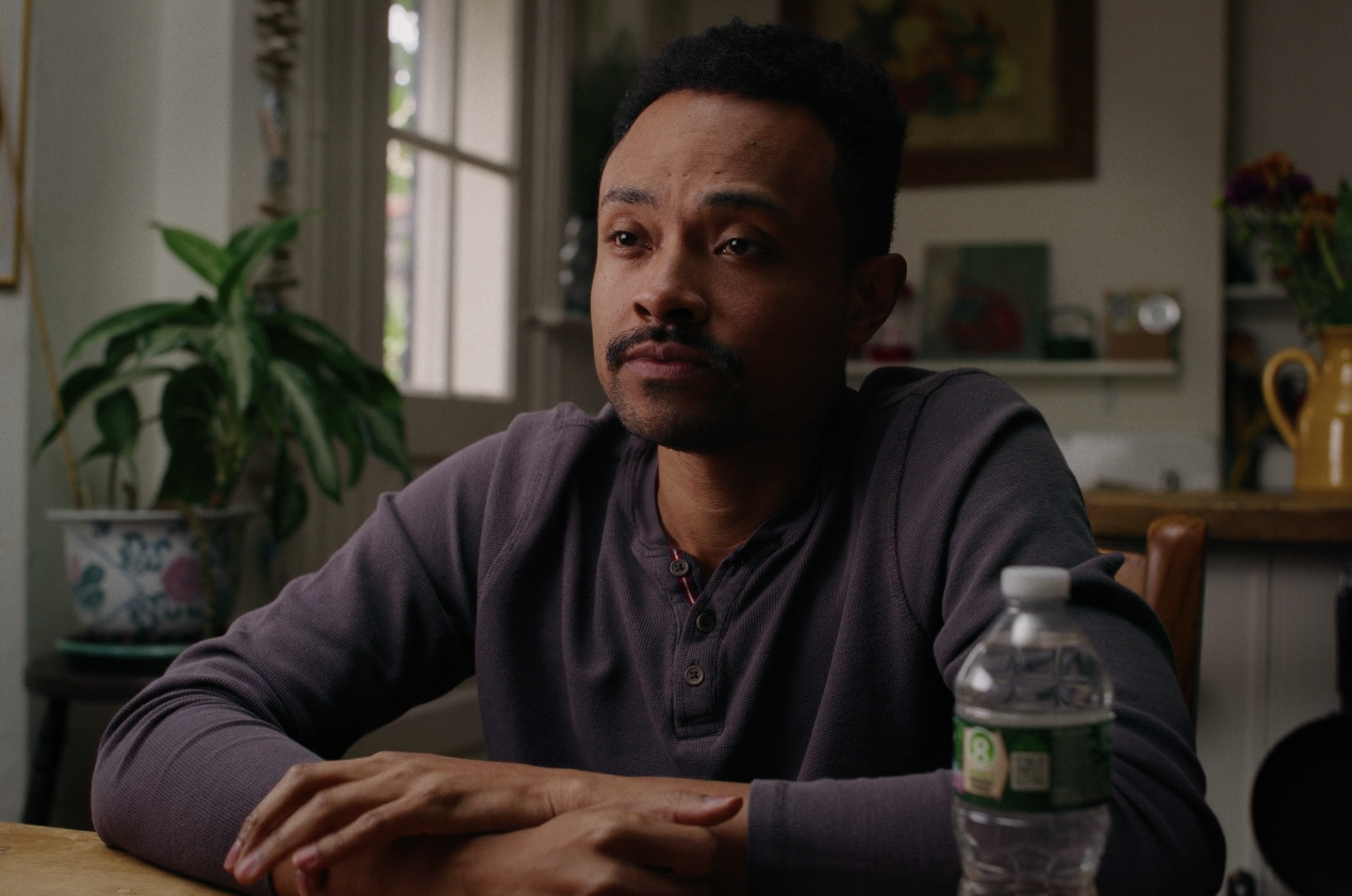 A man with a mustache and short curly hair, sitting at a table in a room with a window, plants, and bookshelf, resting his arms on the table next to a water bottle.