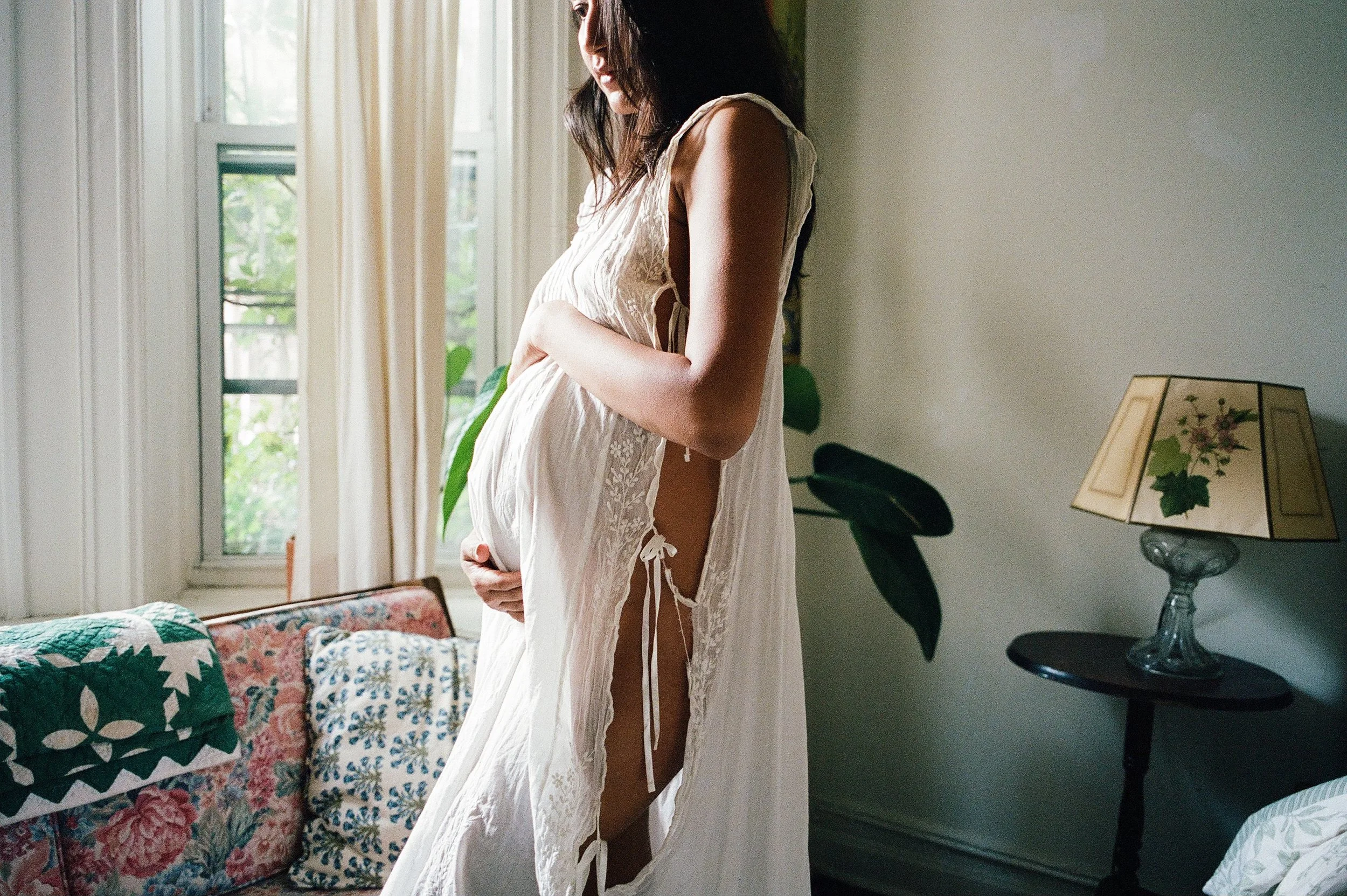 A pregnant woman in a cream-colored lace dress standing in a room near a window, gently touching her belly with both hands, with sunlight coming through the window.