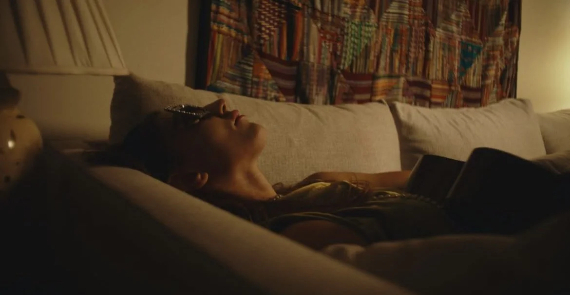 Person lying on a sofa with eyes closed, wearing glasses, in a dimly lit room with a colorful quilt hanging behind.