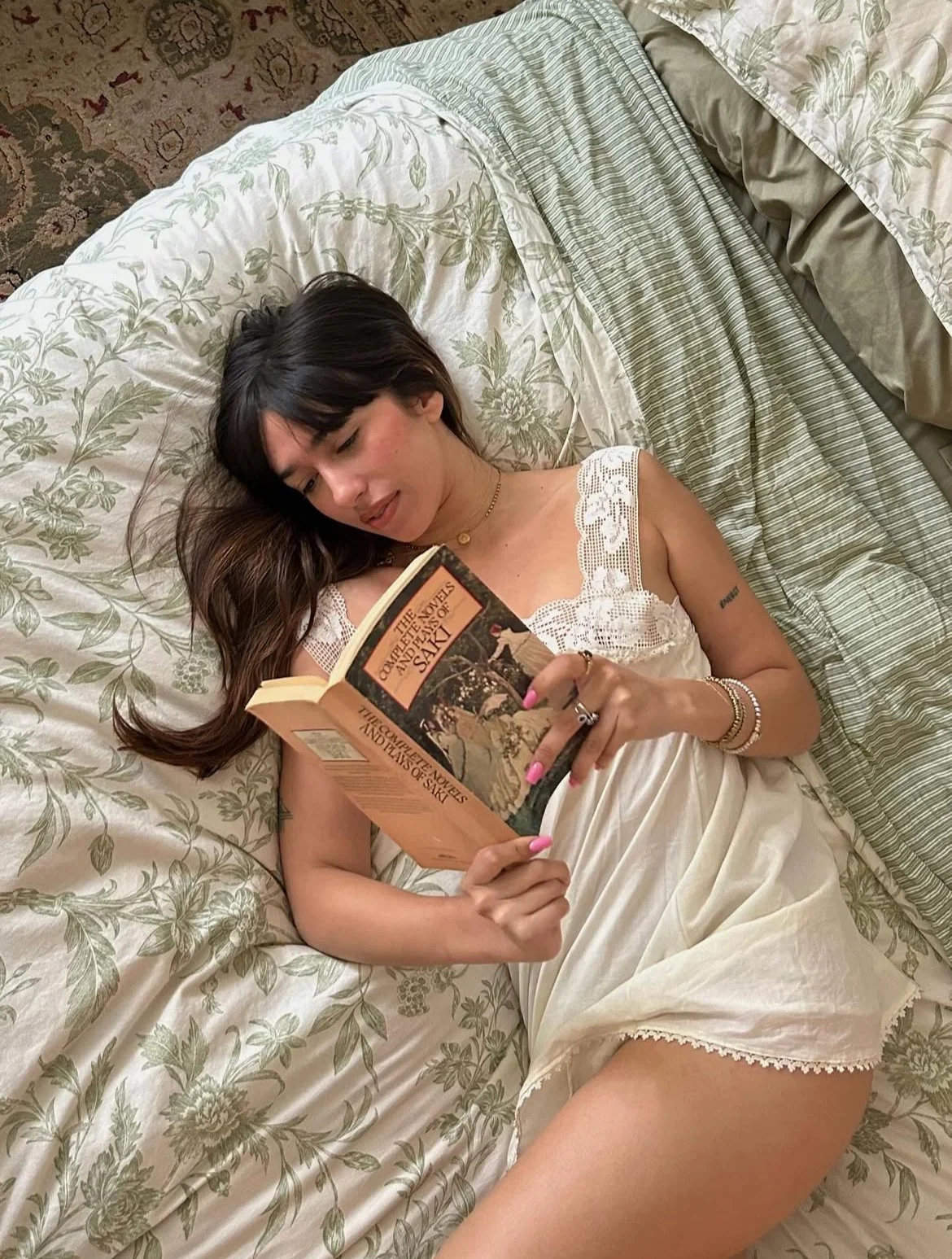 A woman lying on her side on a bed, reading a book titled 'The Complete Novels and Stories of Jane Austen'. She has dark hair with bangs, wears a white nightgown with lace details, and has jewelry including rings and bracelets.