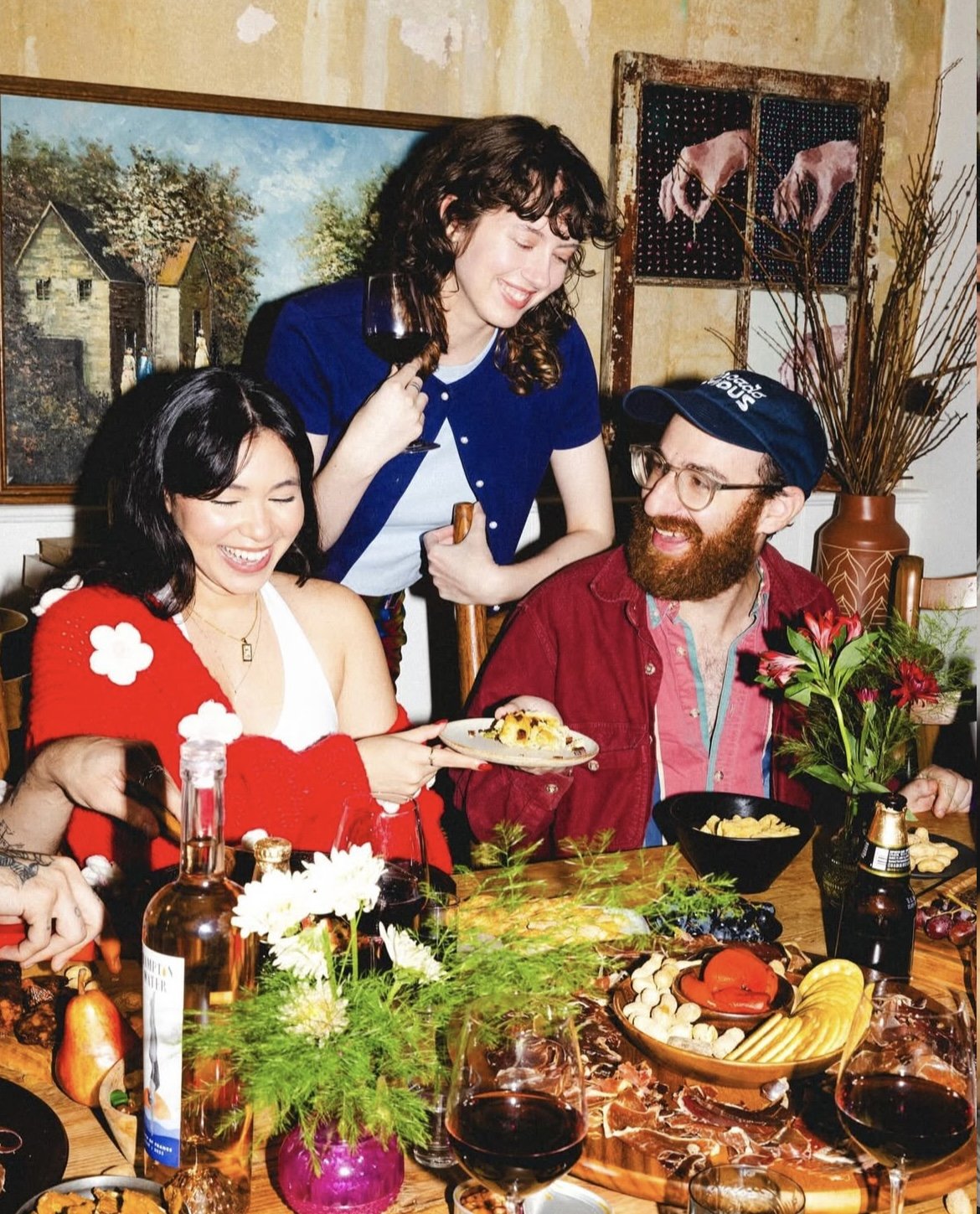 Three people enjoying a meal; a woman in a white tank top with a red shawl, a man with glasses and a beard wearing a red jacket, and a woman with curly hair in a blue shirt holding a glass of wine, all gathered around a table with food and drinks.