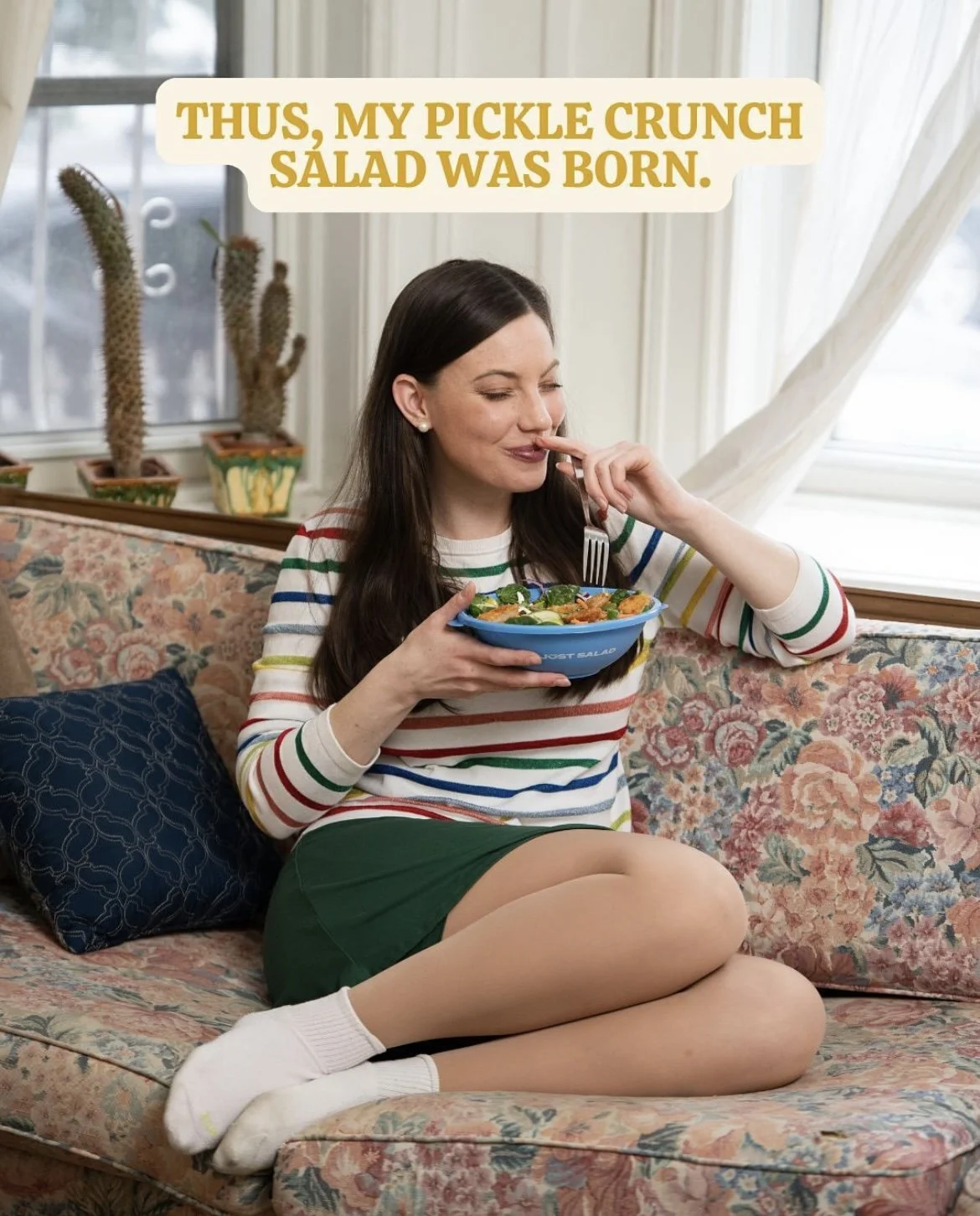 A woman sitting on a floral couch, eating a salad from a blue bowl, wearing a striped shirt, green shorts, and white socks, with cacti on the windowsill behind her, and a speech bubble that says, "Thus, my pickle crunch salad was born."