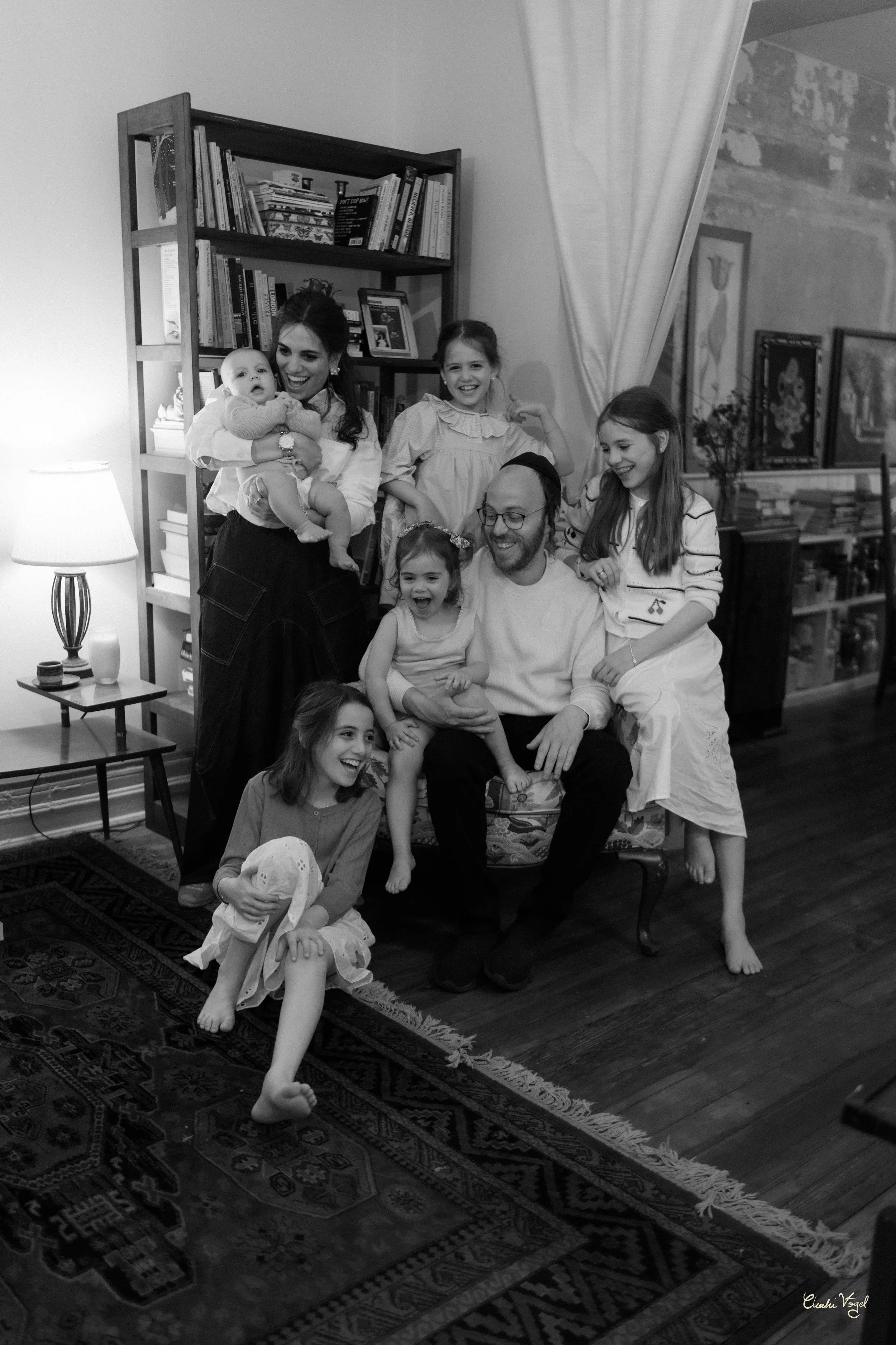 Family gathering in living room, seven children and one adult, some children sitting on adult's lap, others standing or sitting on the floor, all laughing and smiling, with bookshelves, a lamp, and framed pictures in the background.