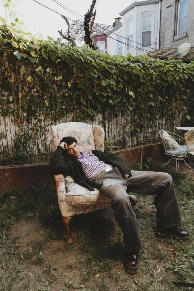A person reclining on a floral upholstered armchair outdoors in a garden, with a vine-covered fence and neighboring houses in the background.