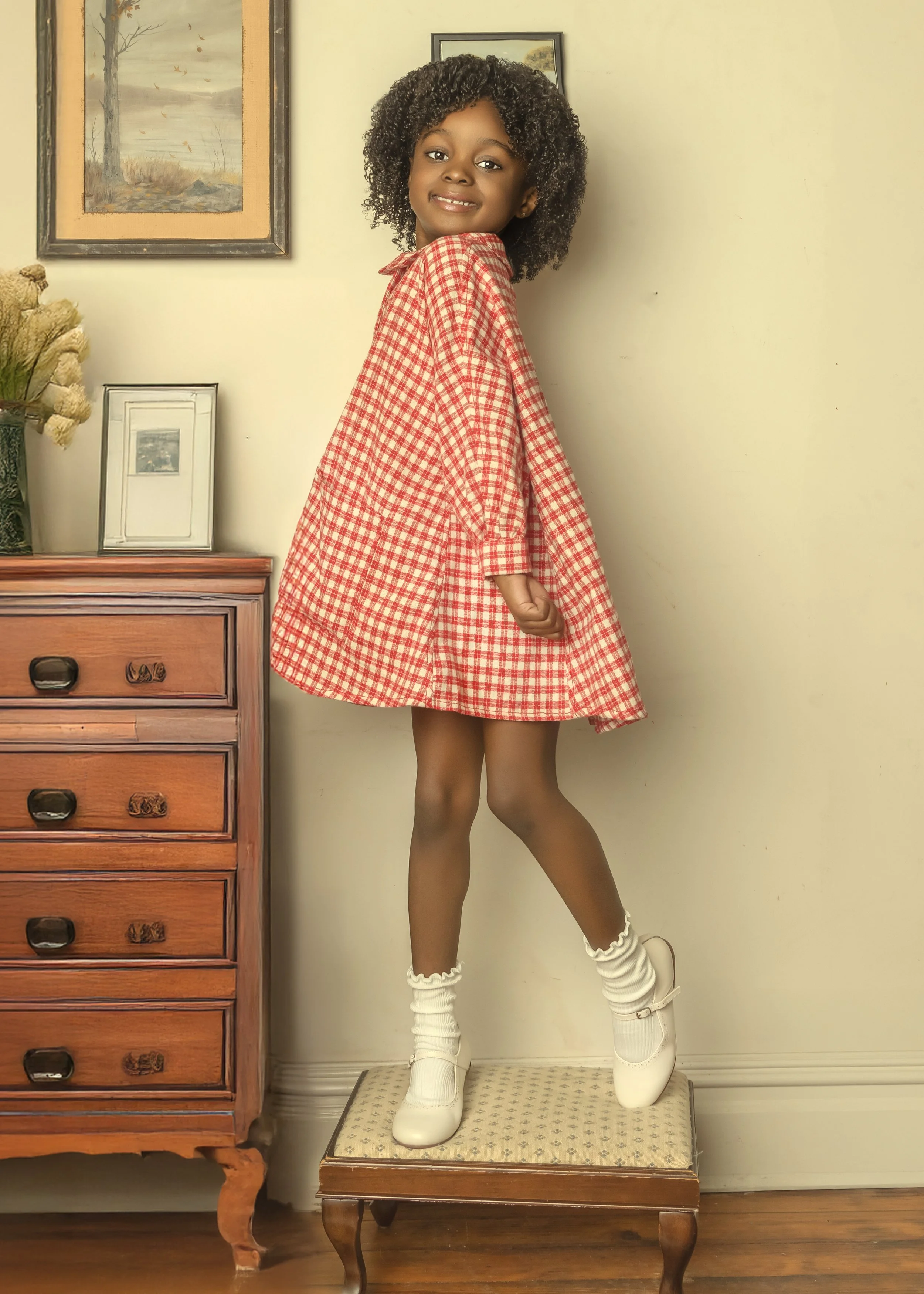 A young girl in a red checkered dress, white socks, and white shoes, standing on a small stool, smiling and posing in a home interior with a wooden dresser and framed pictures on the wall.