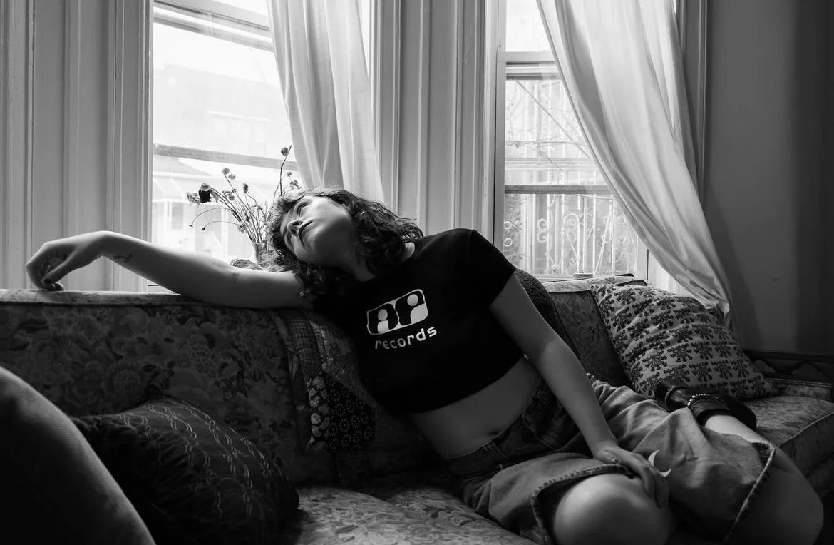 A woman with curly hair wearing a black t-shirt and shorts sitting on a vintage floral sofa, relaxing with her eyes closed near a window with curtains.