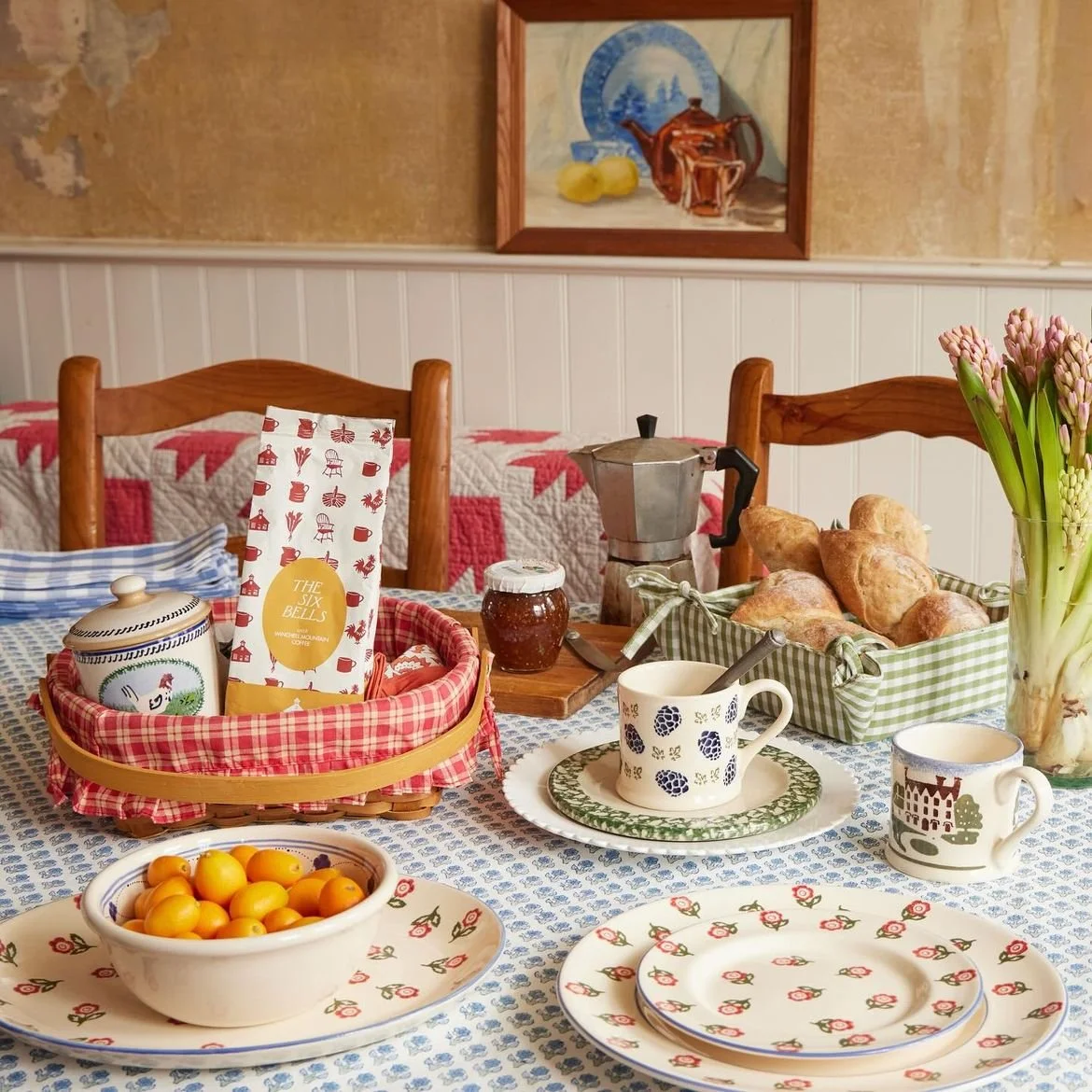 A cozy breakfast table with plates, cups, and a basket of bread in a rustic kitchen or dining room.