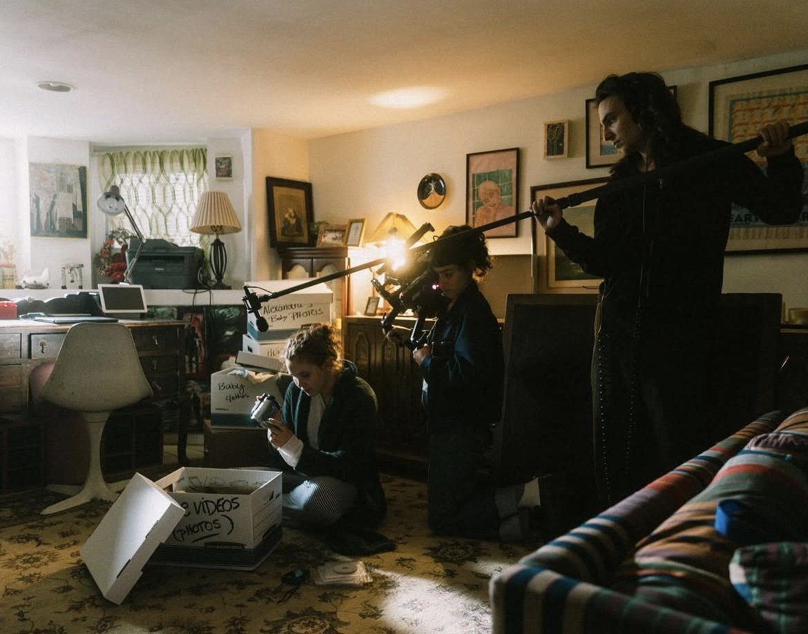 Three women working on a film set inside a living room, with equipment like a camera crane and boxes labeled for videos and photos.