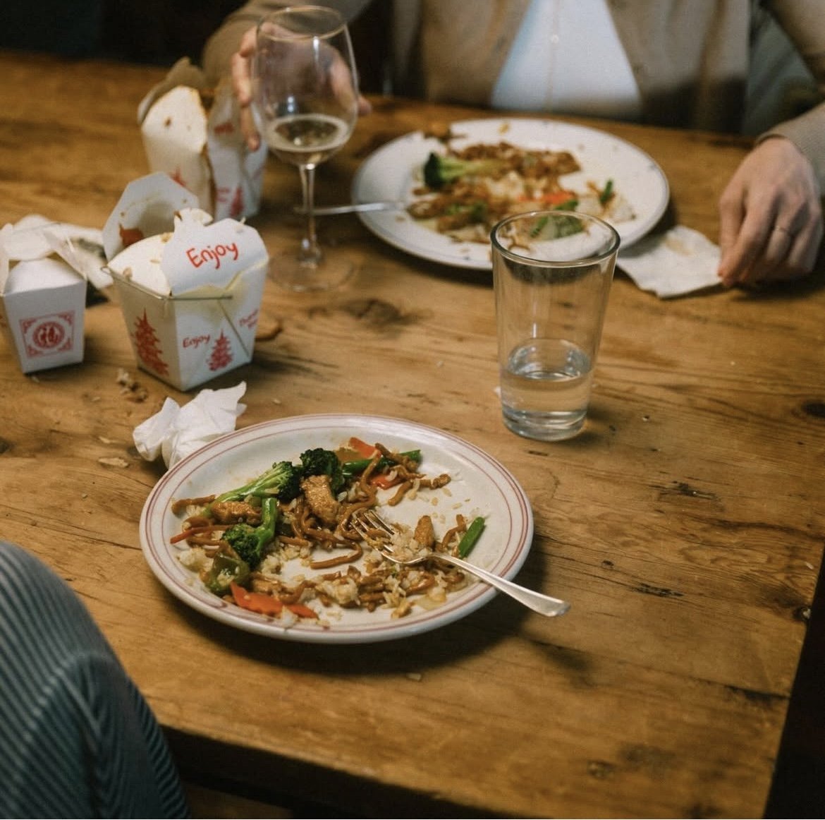 Partially eaten plate of stir-fried vegetables and rice, empty snack containers, a glass of water, and a glass of white wine on a wooden table with people in the background.