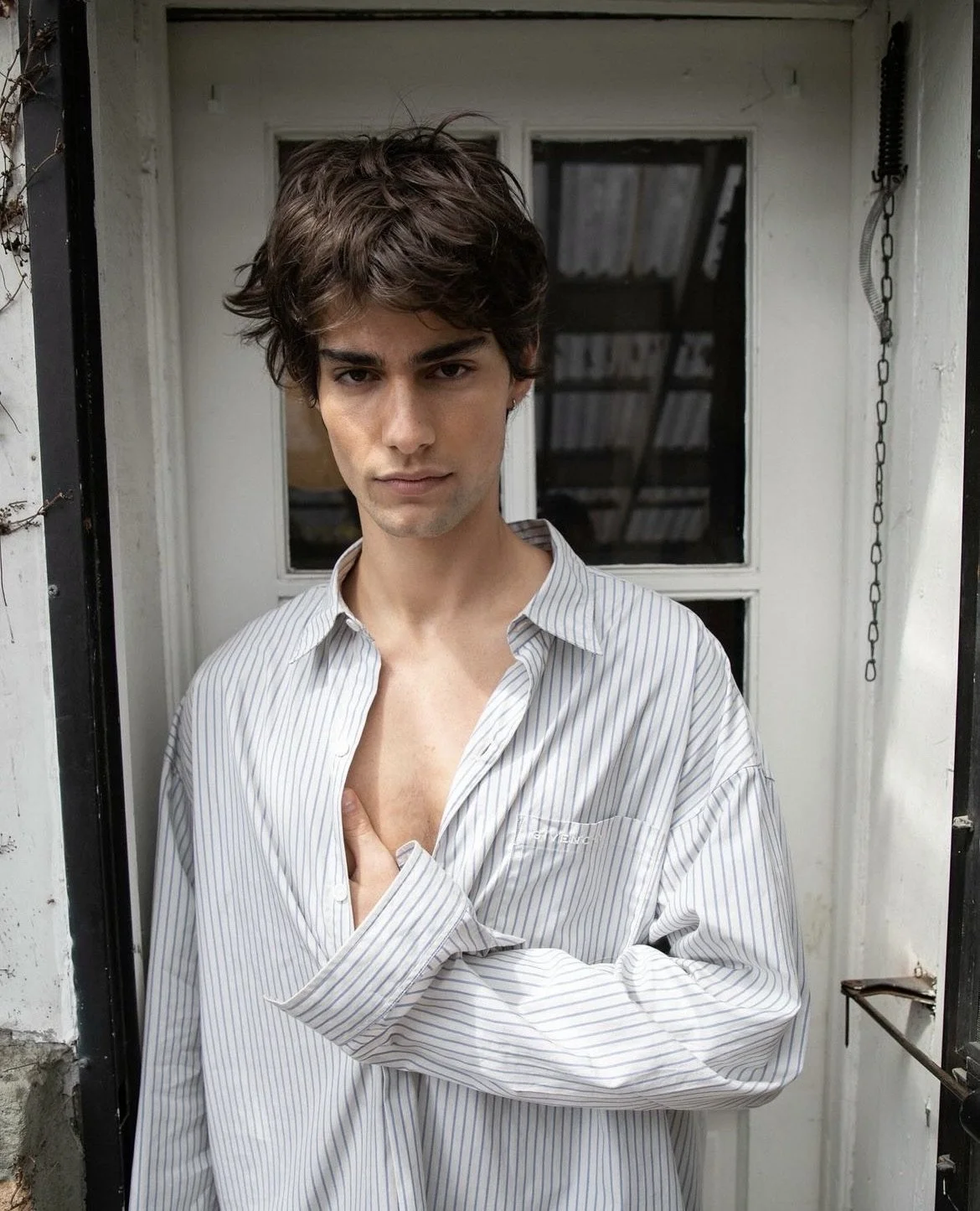 A young man with dark, curly hair and light skin standing outdoors in front of a white door with a window, wearing a striped button-up shirt slightly open to reveal part of his chest, looking directly at the camera.