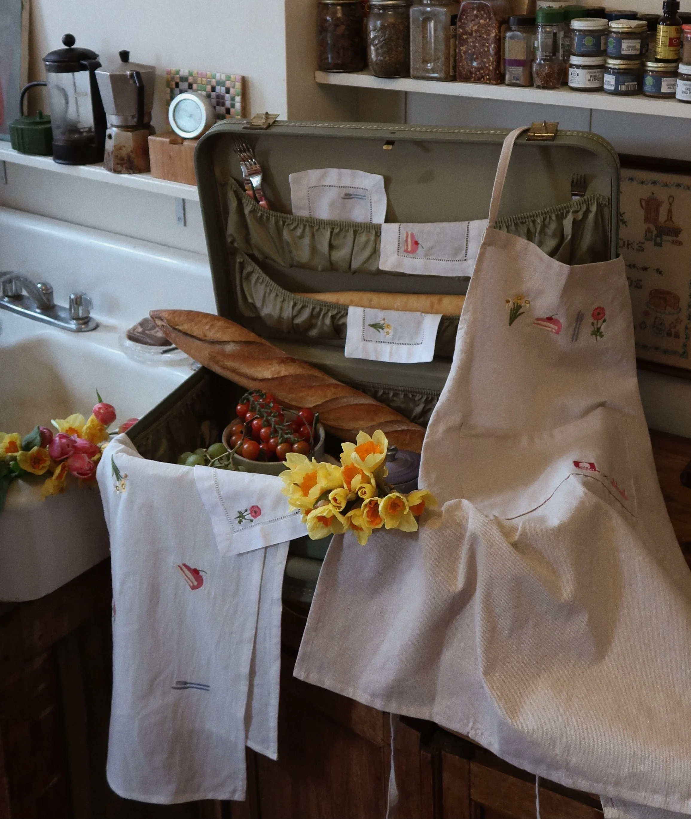 Open vintage suitcase with a baguette, cherry tomatoes, and grapes inside, decorated with floral cloth and artificial flowers, placed on a kitchen counter beside a sink and spices on shelves.