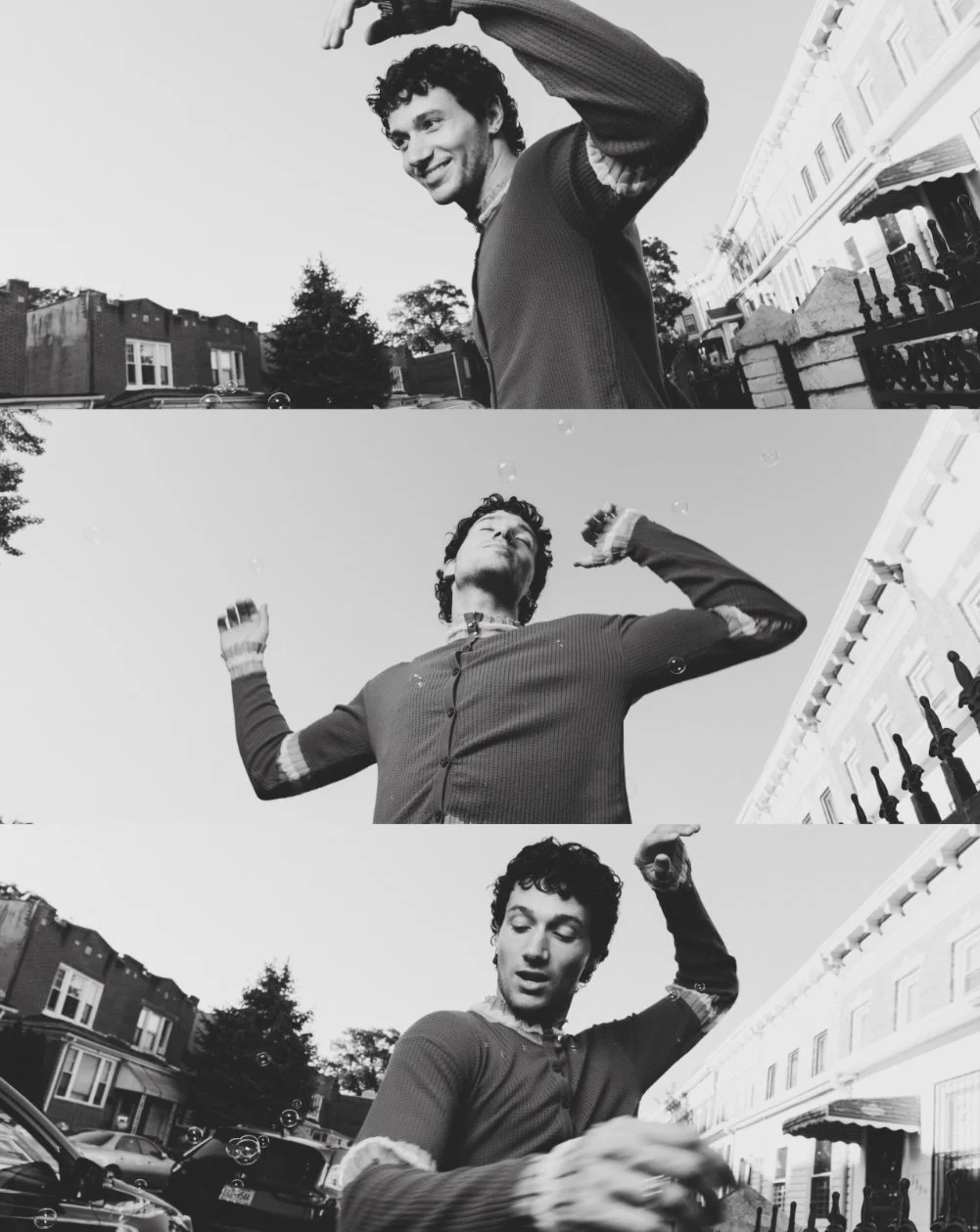 A black and white photo series of a young man with curly hair, wearing a long-sleeve shirt, posing outdoors on a city street with buildings, parked cars, and a fence in the background. In the first image, he is smiling and waving; in the second, he i