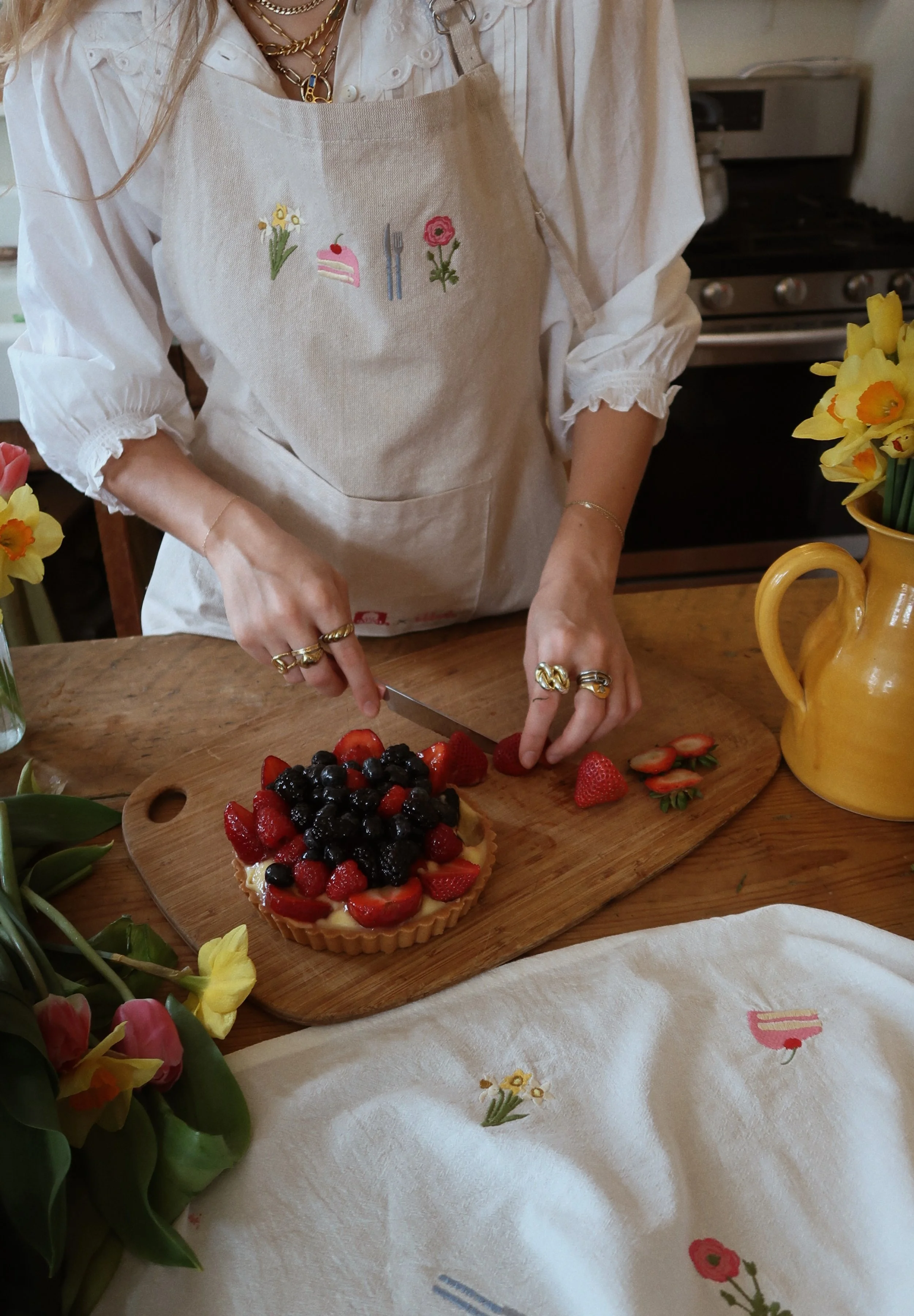 Person slicing strawberries on a wooden cutting board next to a berry tart with fresh berries, wearing a beige apron with embroidered flowers, a white blouse, and gold jewelry, in a kitchen with yellow flowers on the counter.