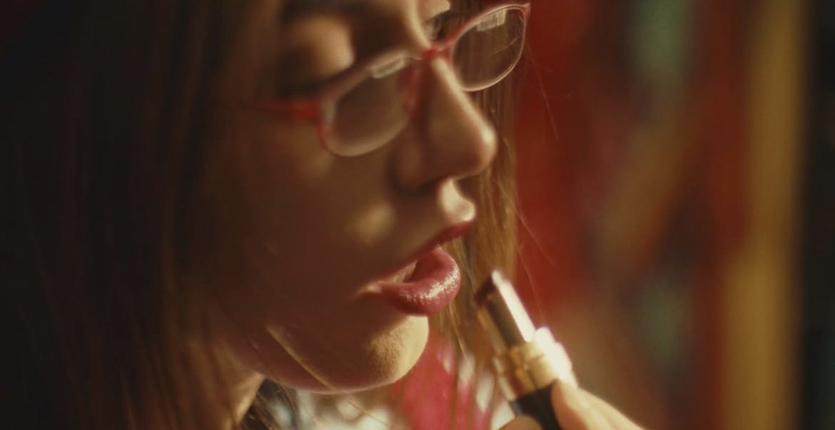 A woman with red hair and glasses applying lipstick.