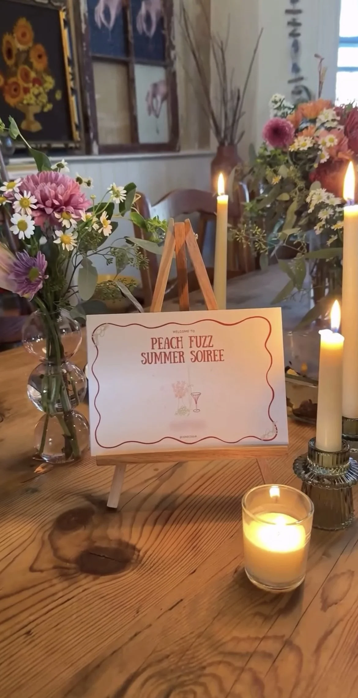 A table decorated for a summer soirée with a small sign, flowers in vases, and lit candles.