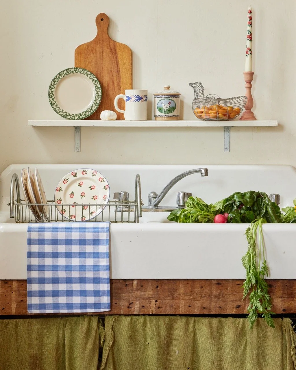 Kitchen sink with a blue and white checkered towel hanging on the side, metal dish rack with plates, green leafy vegetables, and radishes inside the sink. Shelves above with a cutting board, plates, a mug, a decorative tin, a wire basket shaped like 