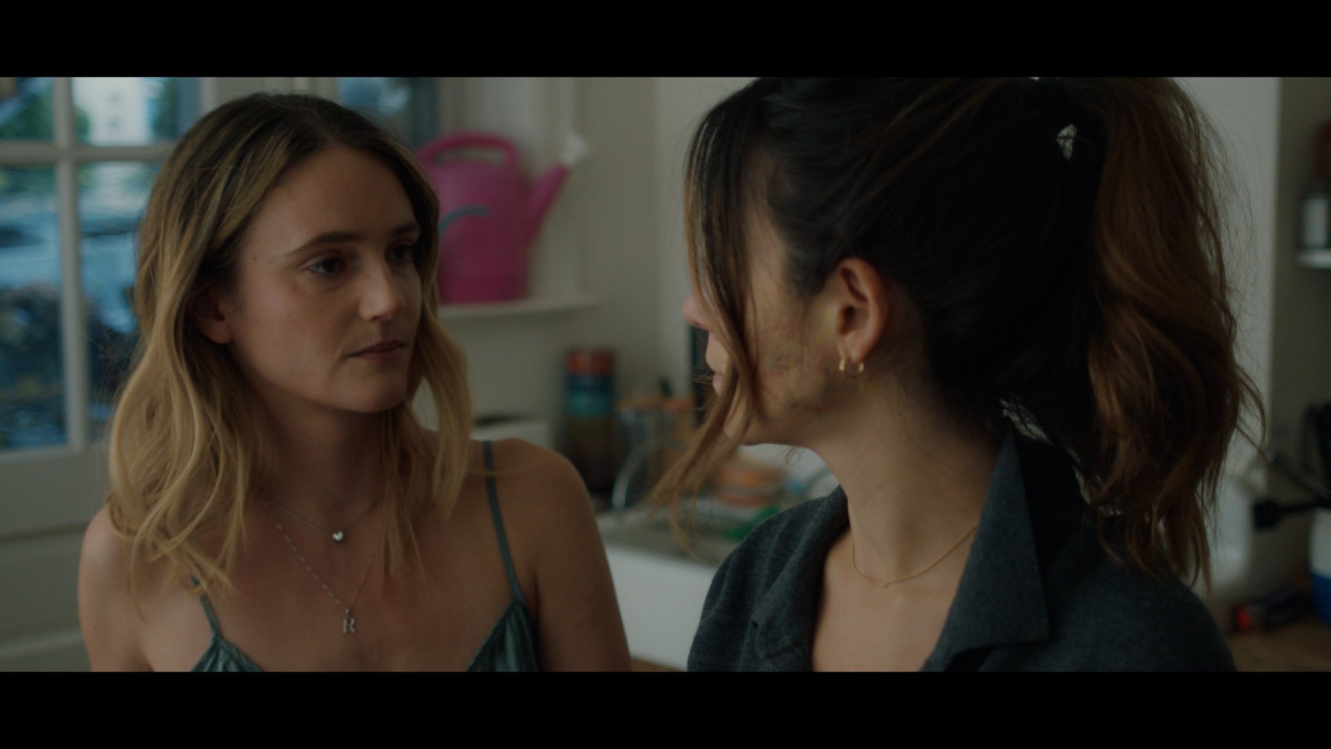 Two women face each other in a kitchen, one with blonde hair and the other with dark hair tied back, engaged in a serious conversation.