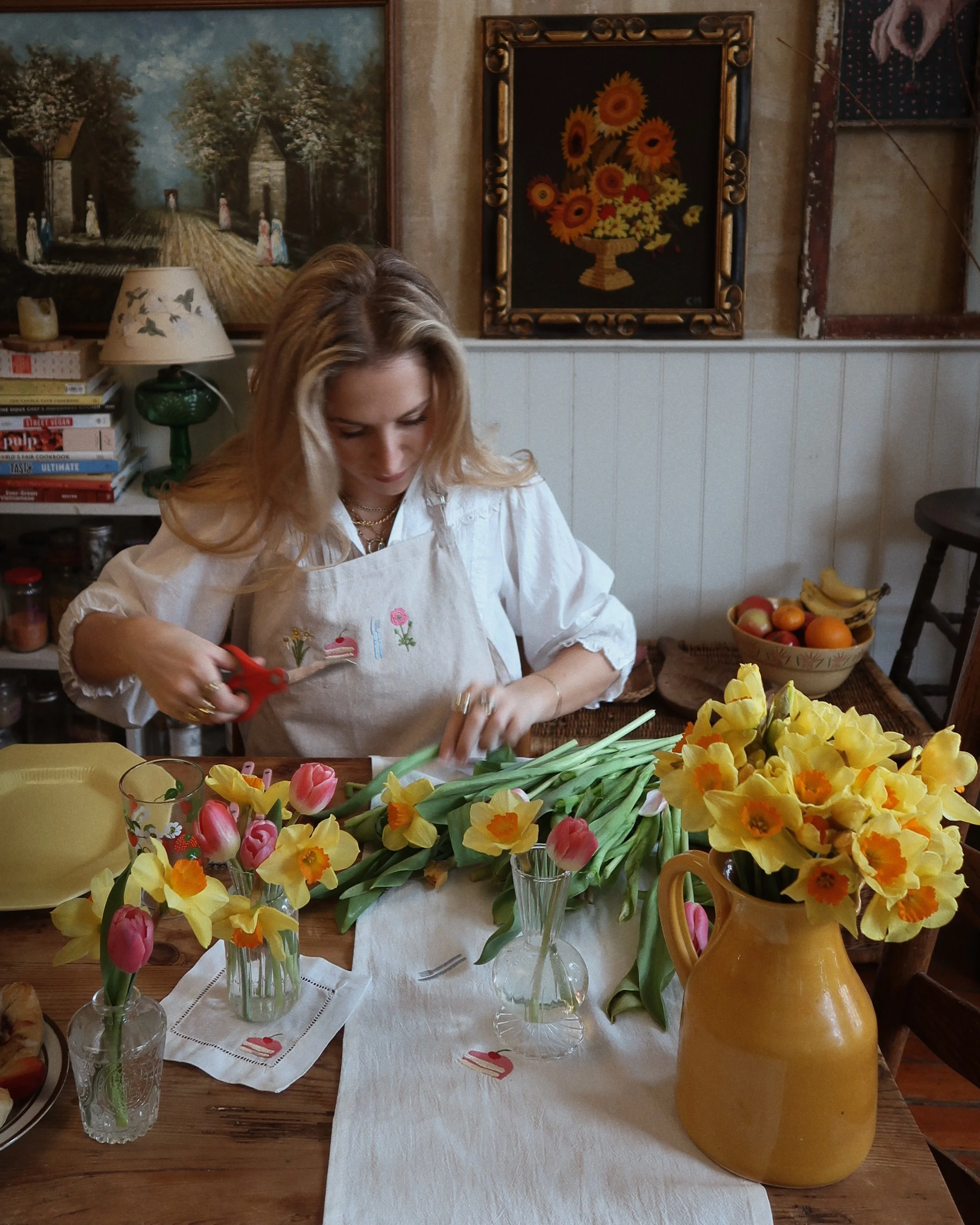 A woman wearing a white apron is cutting tulips and arranging flowers on a table with vases and a large yellow jug filled with yellow and pink flowers in a cozy, old-fashioned kitchen or dining room.