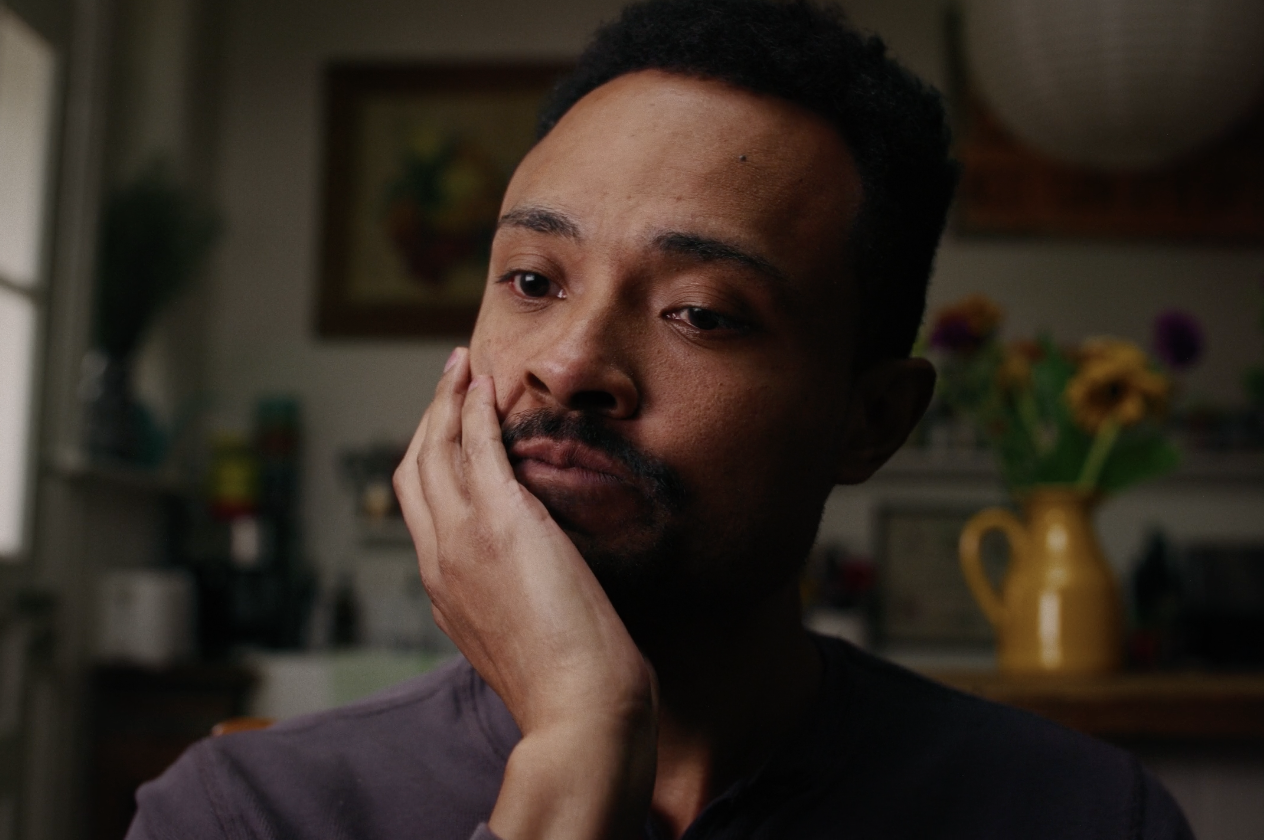 A man with short curly hair and a mustache, resting his face on his hand, appears to be deep in thought in a cozy room with a yellow vase with flowers in the background.