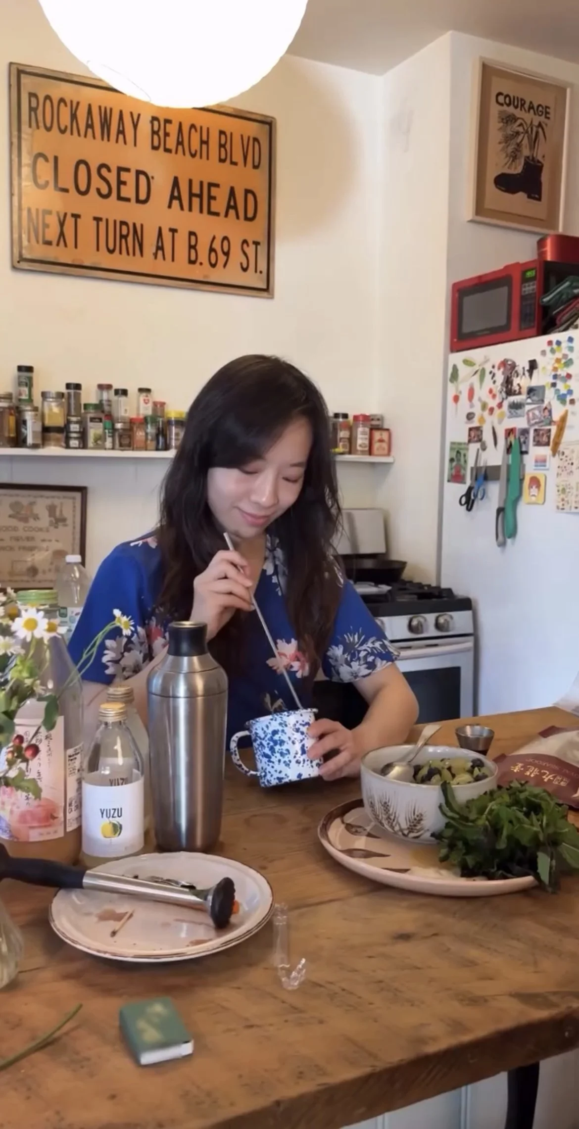 A woman sitting at a wooden table in a kitchen, drinking from a patterned mug, with various condiments and dishes on the table. The background shows shelves with spices, framed art, a stove, and a refrigerator covered with magnets and photos.
