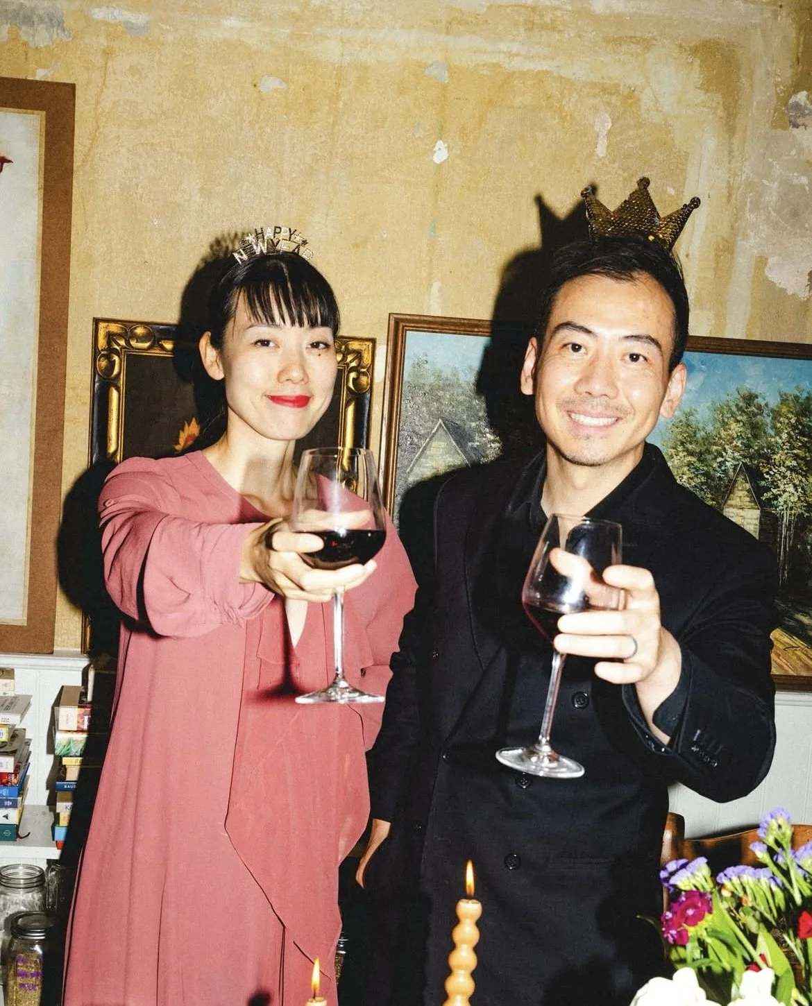 A woman and a man celebrating with glasses of red wine, both wearing crowns, in a room decorated with paintings and flowers.