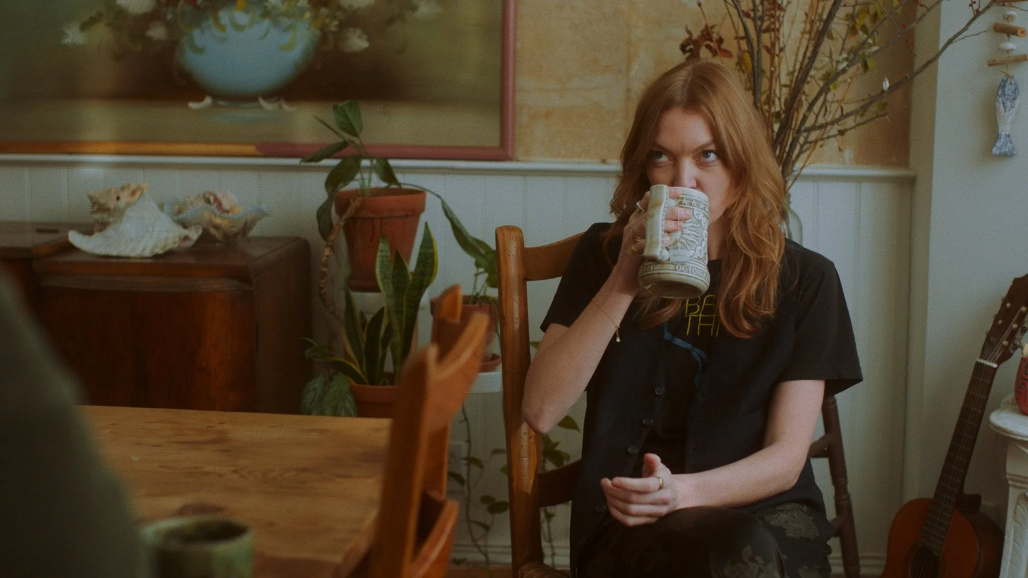 A woman with red hair sitting at a wooden table, drinking from a mug in a cozy room decorated with plants, shells, and a guitar.