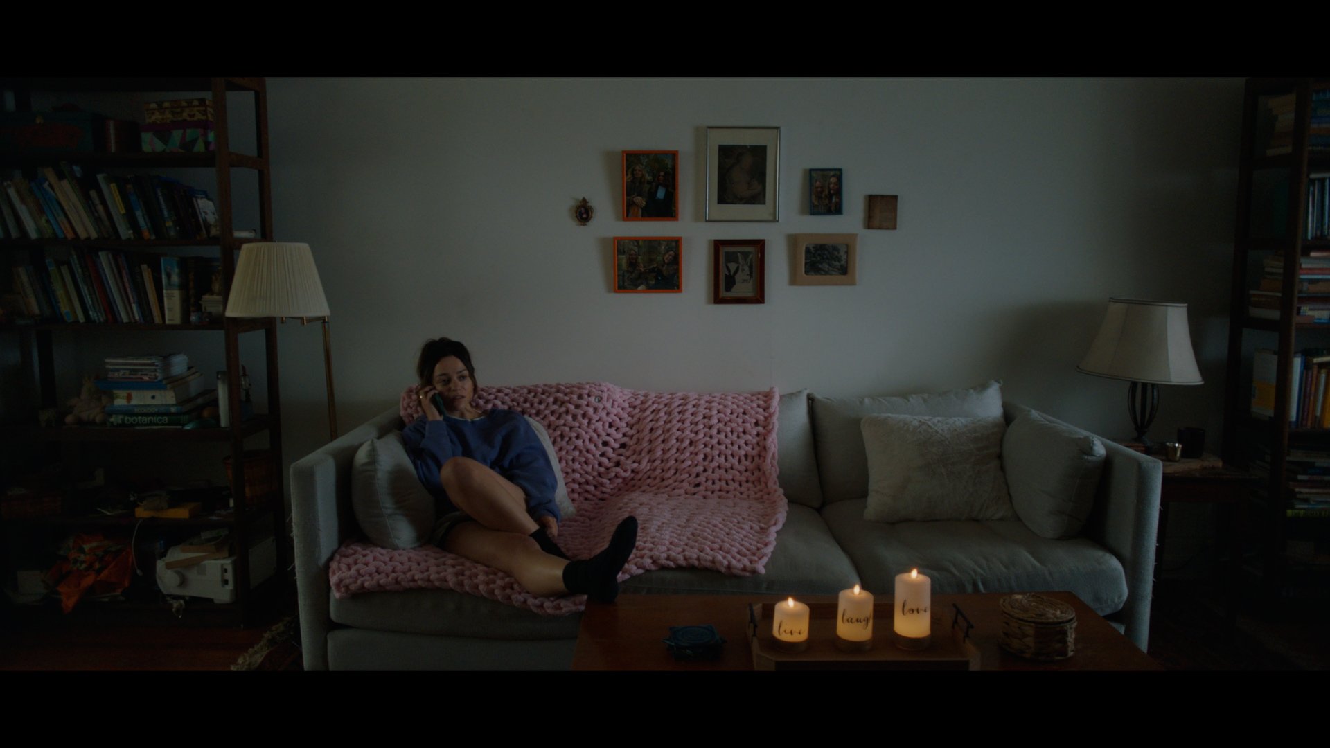 A woman sitting on a beige sofa talking on the phone in a dimly lit living room, with a pink blanket on her lap and shelves filled with books, framed pictures on the wall, and candles on a table.