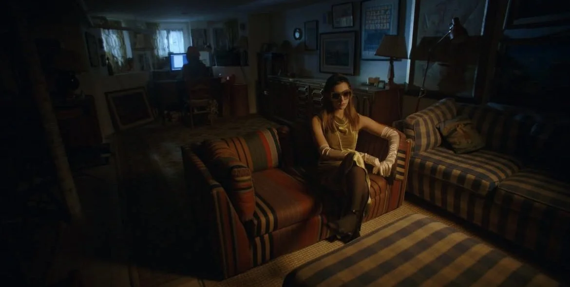 A woman wearing sunglasses, a dress, and gloves sitting on a striped sofa in a dimly lit room, with a man working on a computer in the background.