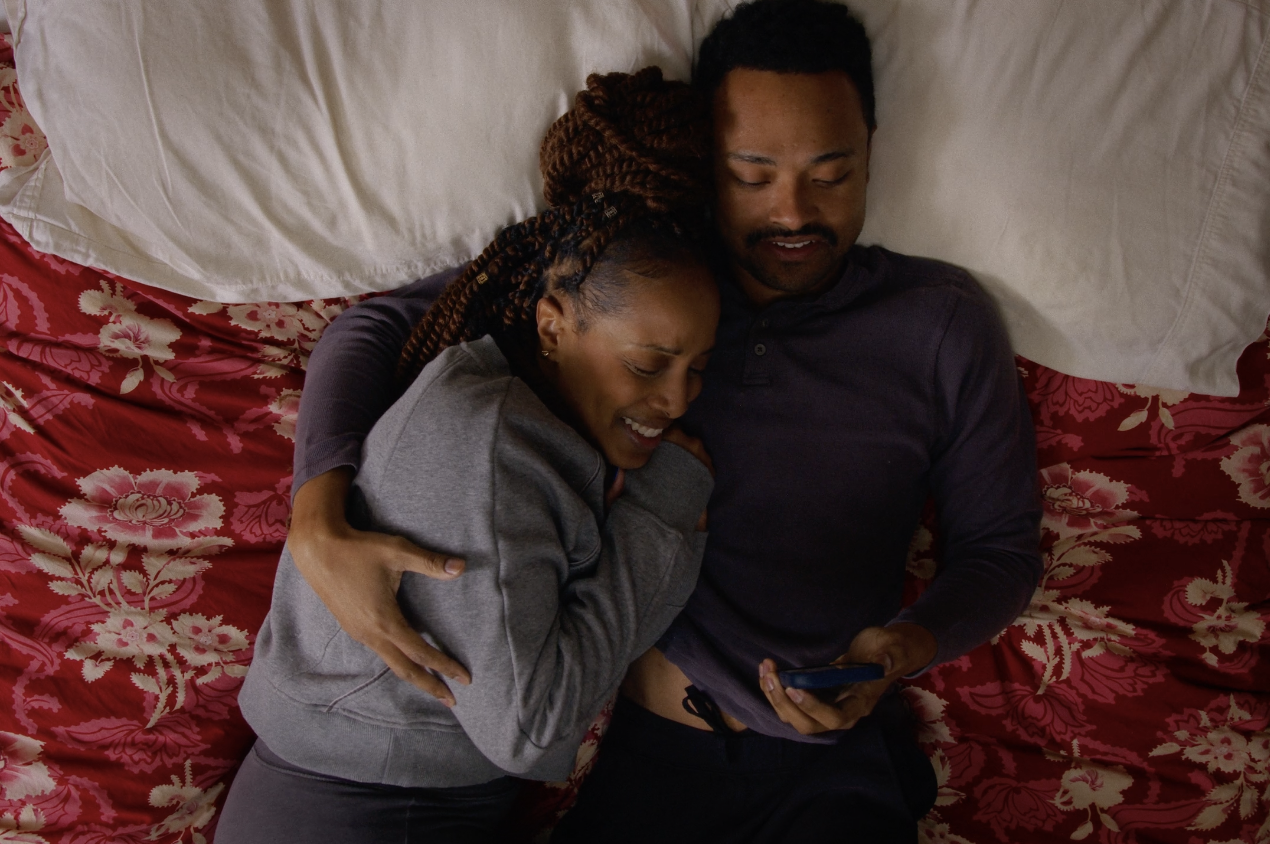 A couple lying in bed together, laughing and looking at a phone.
