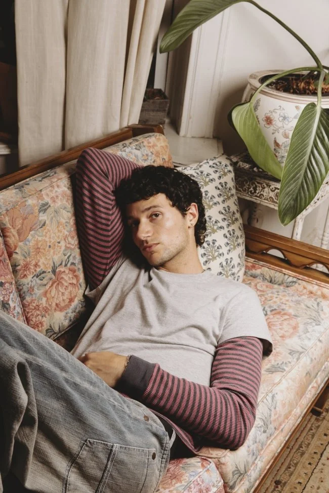 A young man with dark curly hair reclining on a floral-patterned couch with his left arm behind his head, wearing a gray T-shirt over a long-sleeve red and black striped shirt, and gray pants, in a room with beige curtains, a large green houseplant, 