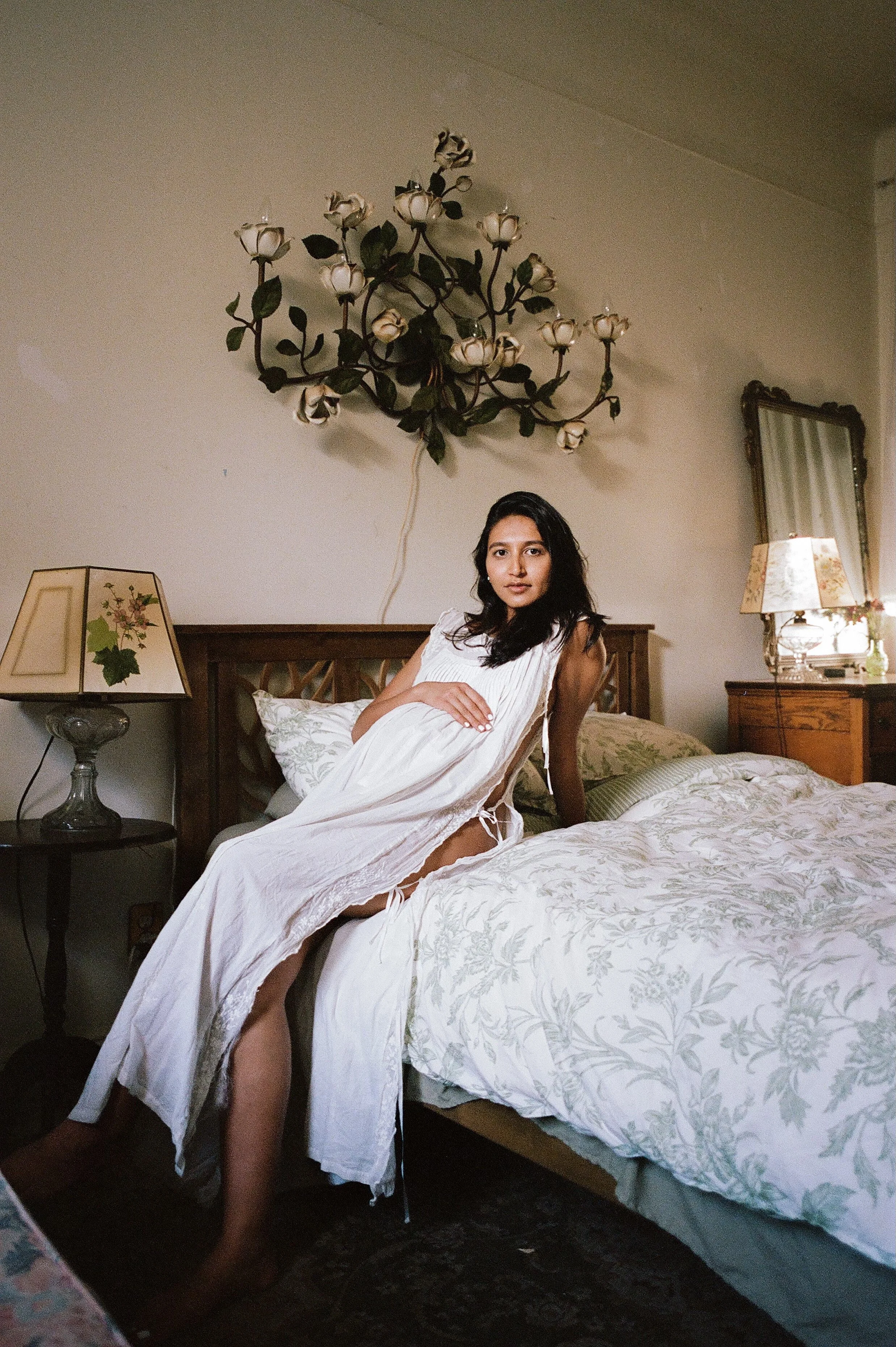 A young woman in a white dress sitting on a bed in a bedroom, with lamps and floral decorations visible in the background.