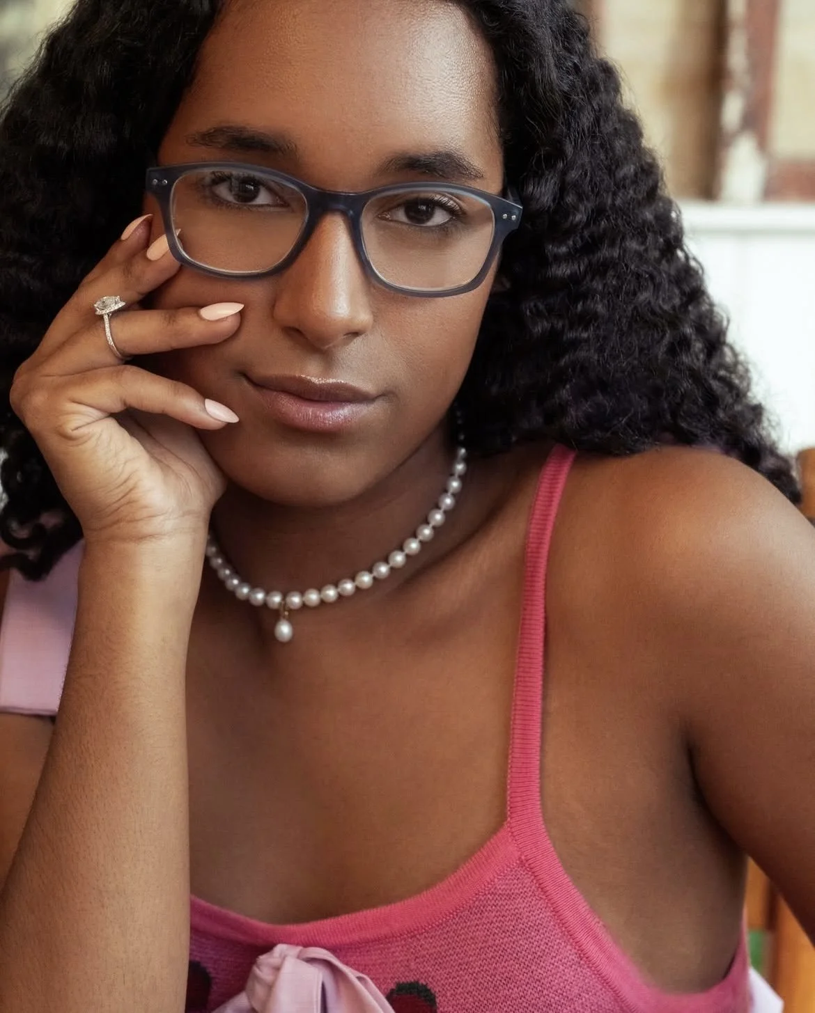 A woman with curly hair wearing glasses, a pearl necklace, a pink tank top, and a ring, resting her face on her hand.