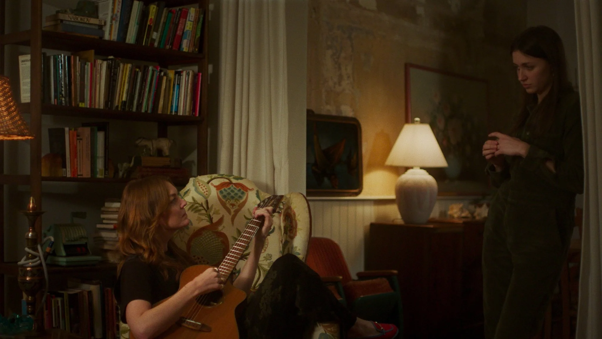 A woman with red hair is sitting on a floral armchair and playing an acoustic guitar while another woman with dark hair is standing nearby, looking down. The scene is in a cozy, warmly lit room with bookshelves and a lamp in the background.