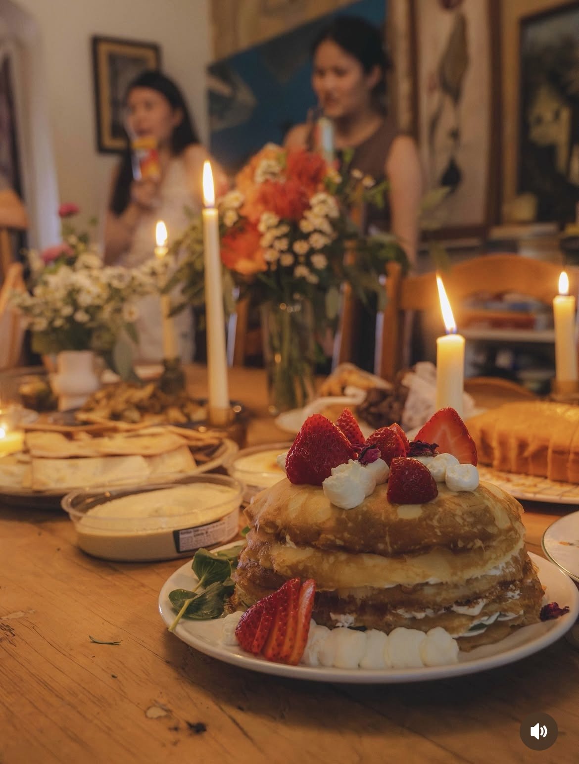 A birthday cake with strawberries and whipped cream on a dinner table, surrounded by candles, with blurred women holding drinks and flowers in the background.