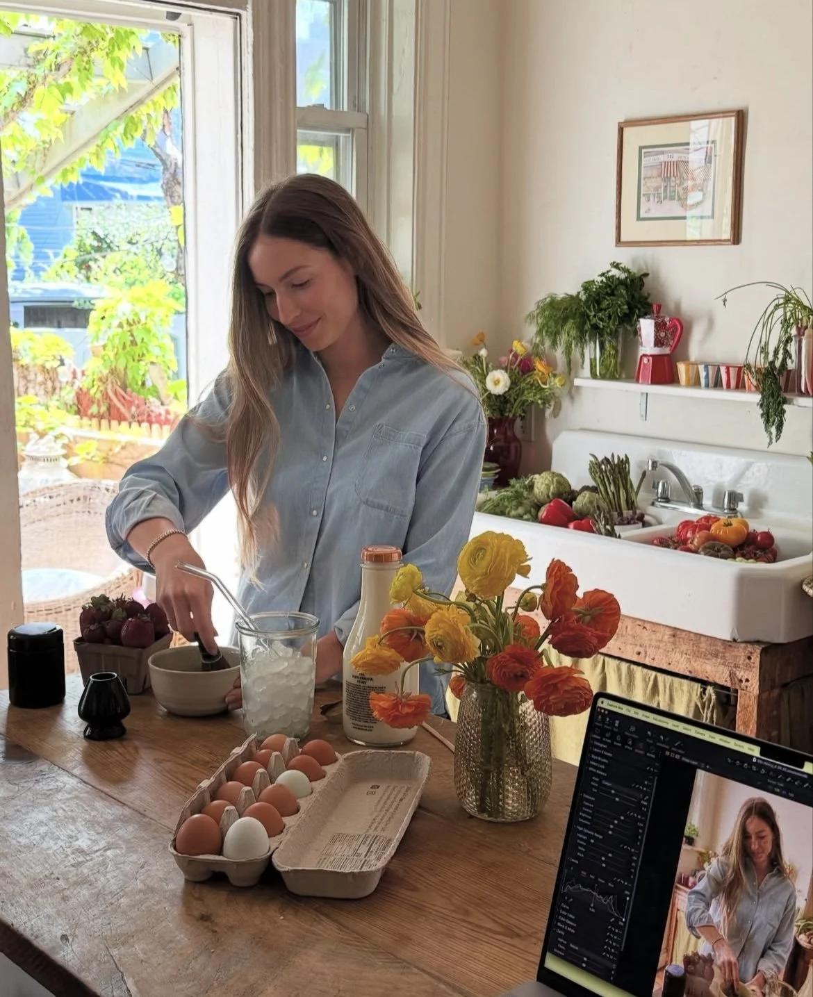 A woman with long hair wearing a light blue button-up shirt, preparing eggs in a kitchen filled with fresh vegetables and flowers, with a laptop capturing her image.