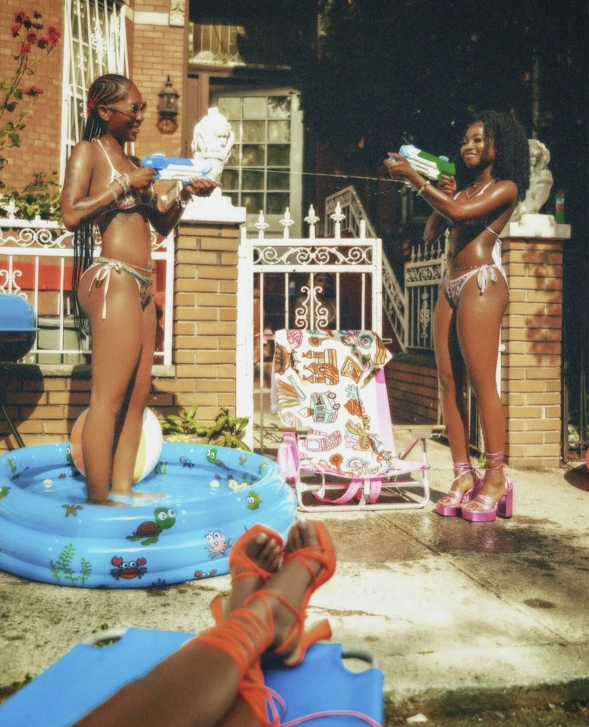 Two women in swimsuits are playing with water guns outside a house. One is in a small inflatable pool, and the other is standing on the ground, both smiling. In the foreground, a person's legs and orange sandals are visible, relaxing on a blue lounge
