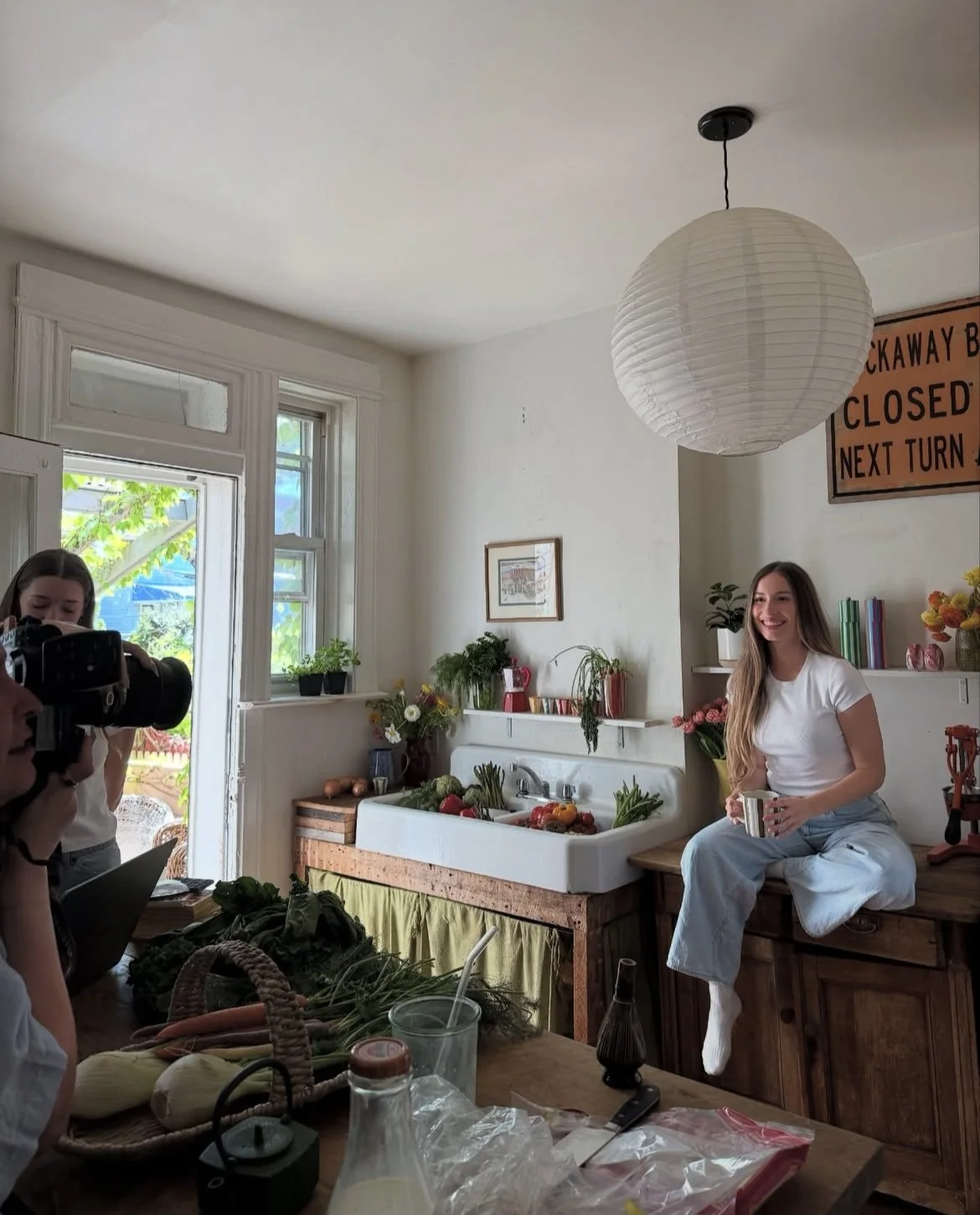 A woman sitting on a kitchen counter holding a mug, smiling, with plants and flowers around her. A person with a camera is taking her picture in a bright kitchen with large windows.