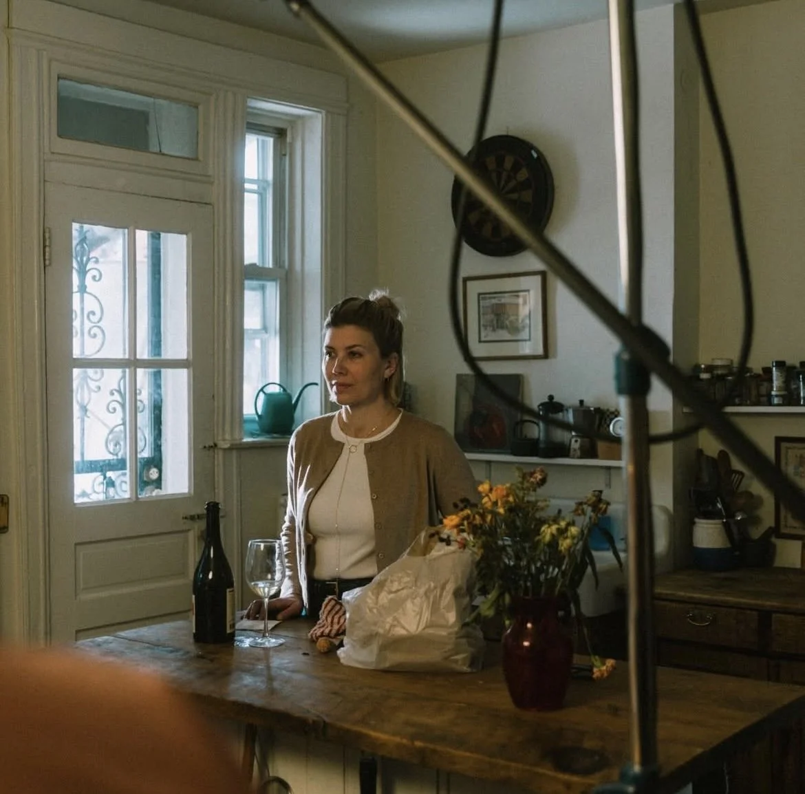 Woman in a beige jacket standing in a cozy kitchen next to a wooden table with wine glass, bottle, and flowers, illuminated by natural light from windows and door.