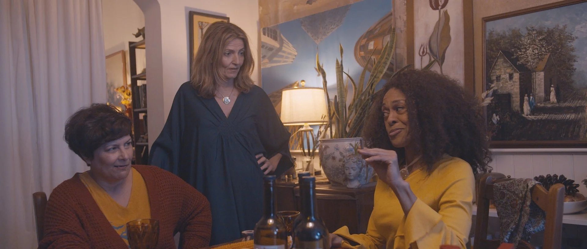 Three women are sitting around a dining table in a warmly lit room, with one woman standing behind them. The woman on the left has short dark hair and is wearing a mustard-colored shirt with a reddish-brown sweater. The woman on the right has curly h