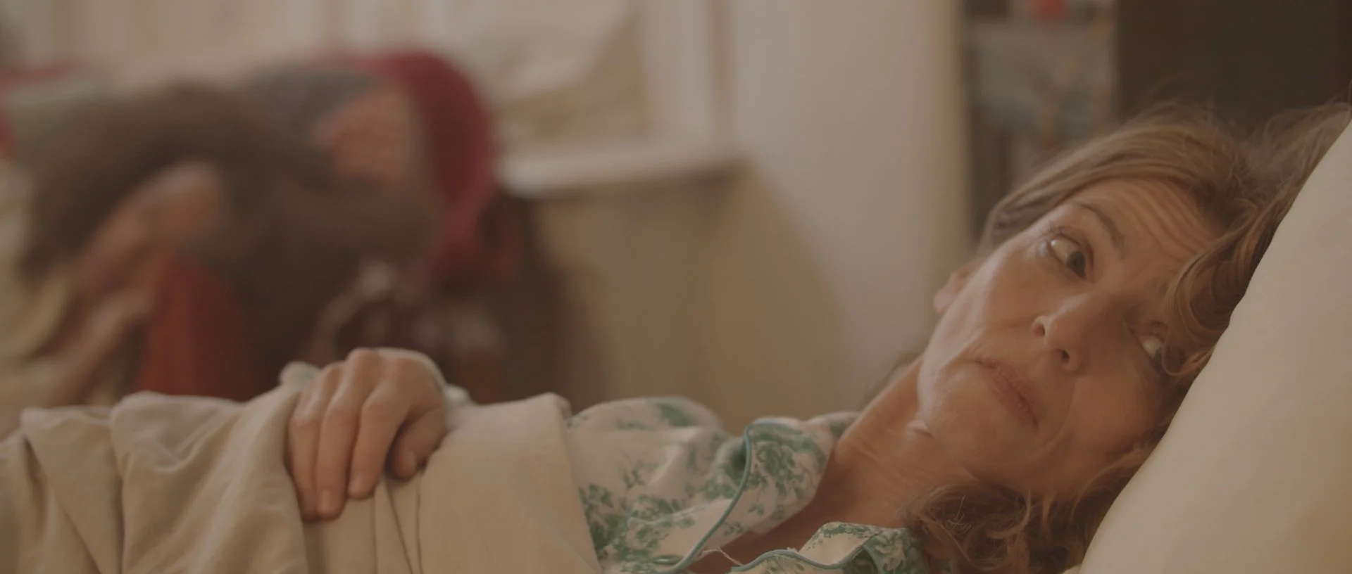 A woman with curly red hair lying in bed, looking worried or contemplative, with her hand resting on her chest.