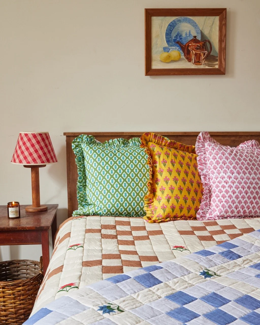 A cozy bedroom with a bed featuring three colorful pillows: green, yellow, and pink, each with ruffled edges. A wooden nightstand with a red plaid lampshade and a lit candle is next to the bed. There is a wicker basket beneath the nightstand. A frame