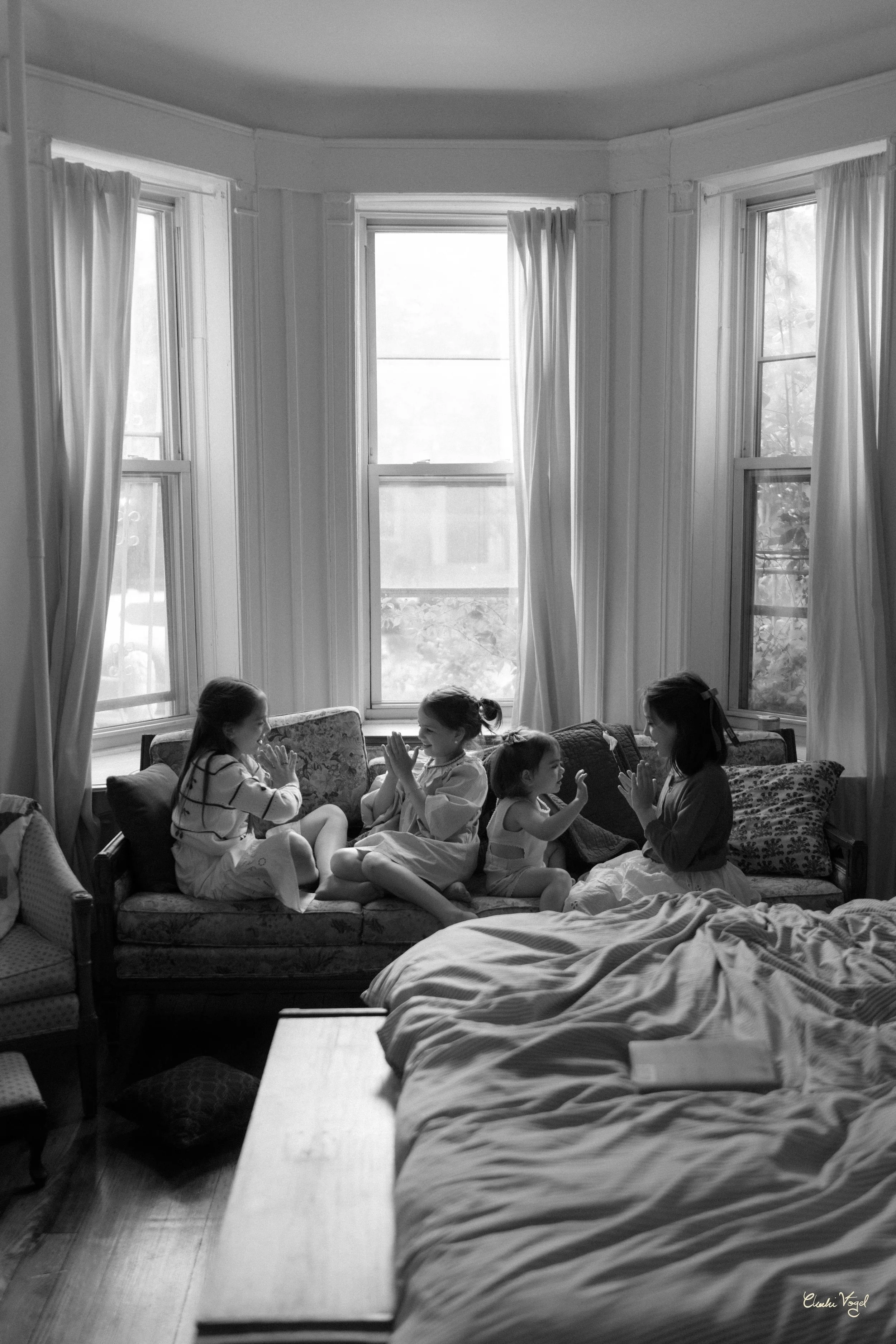 Four girls sitting on a window seat in a bright room, playing a hand game, with curtains, windows, and a bed visible in the background.