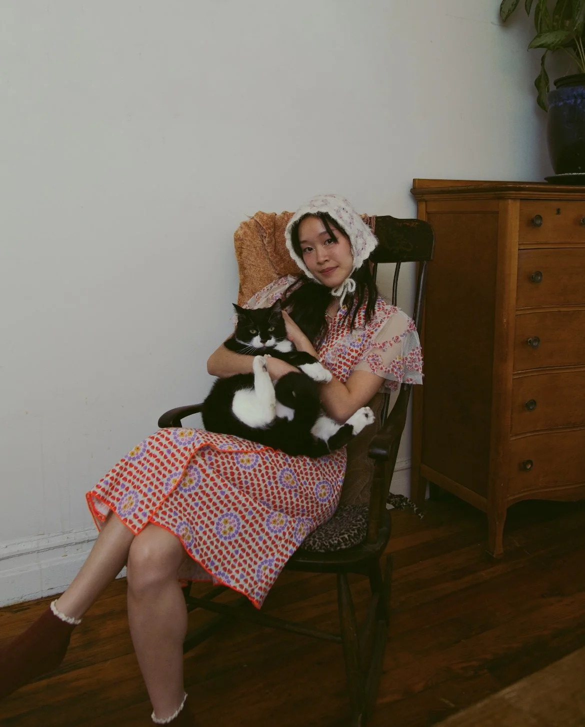 A woman sitting on a chair holding a black and white cat, wearing a floral dress and a cozy hat, in a room with wooden furniture and a potted plant.