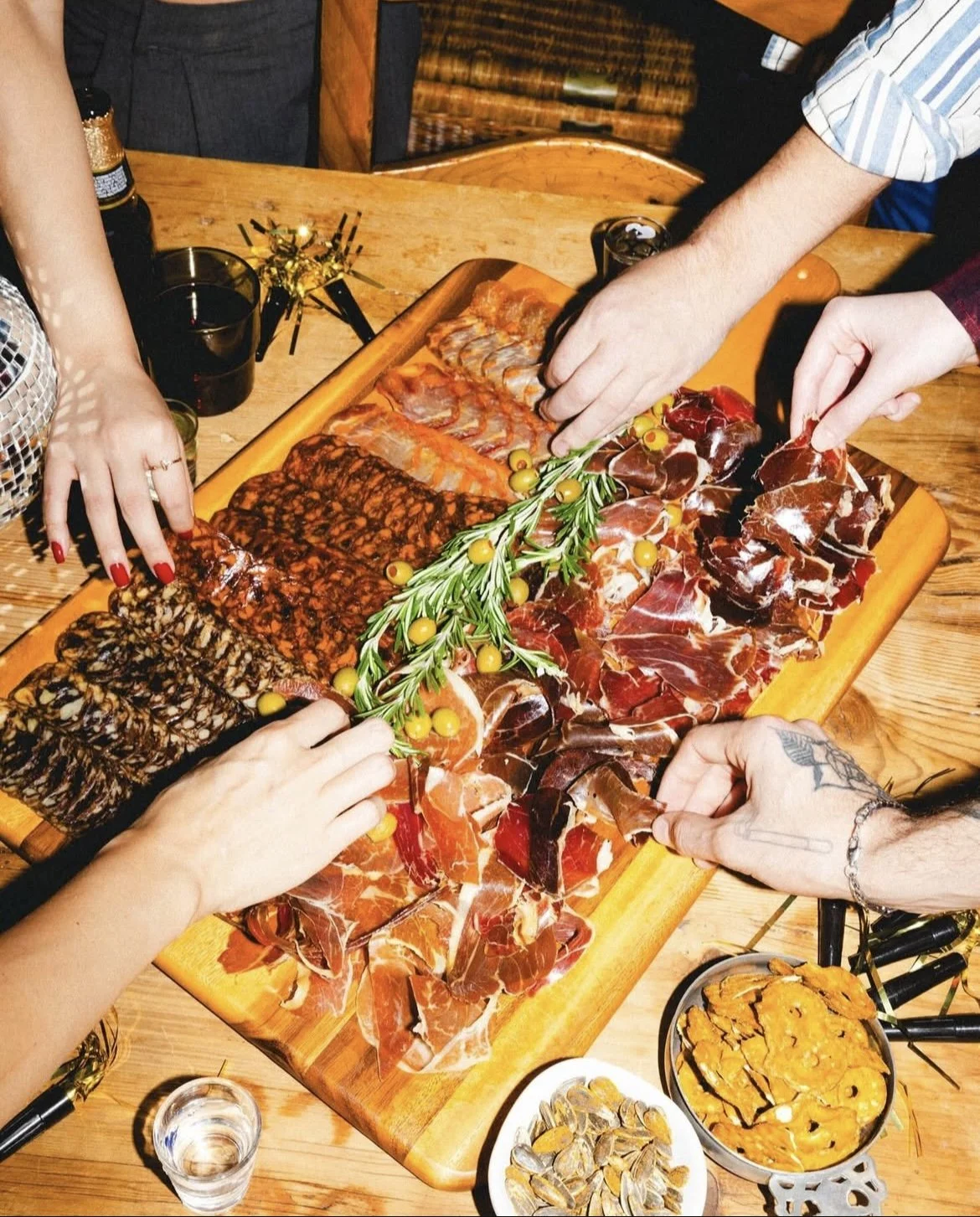 People sharing a platter of assorted cured meats, garnished with sprigs of rosemary and yellow berries at a wooden table along with glasses of drinks and side dishes.