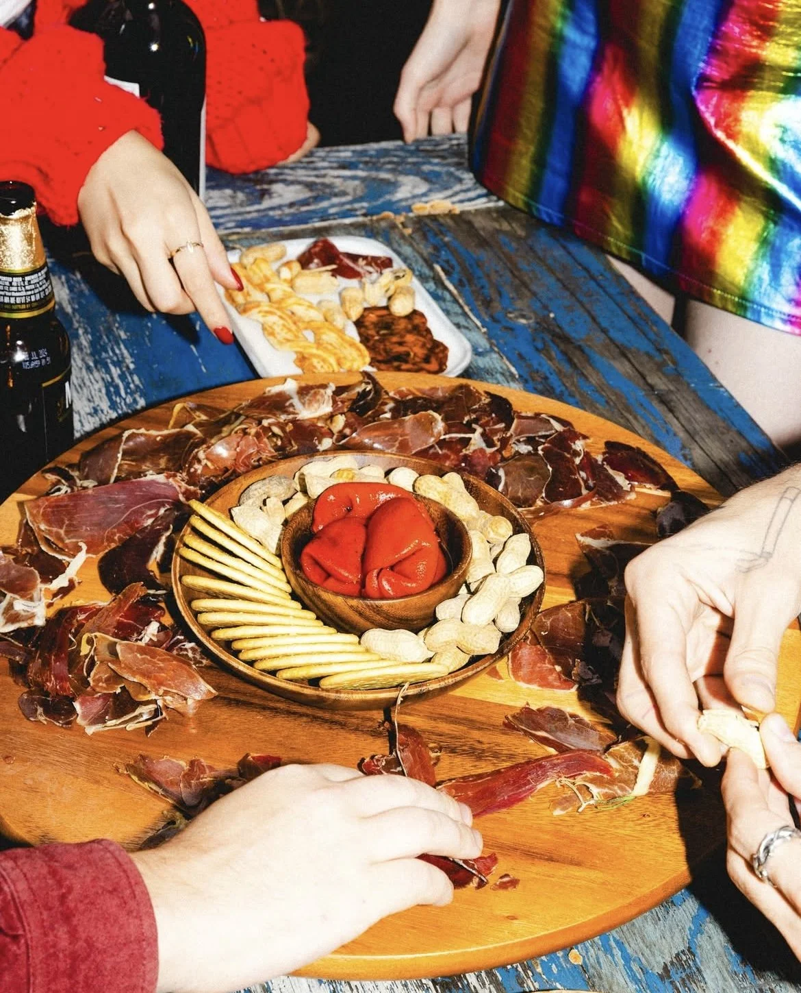 People sharing a charcuterie board with cured meats, cheese, and roasted peppers at a rustic wooden table.
