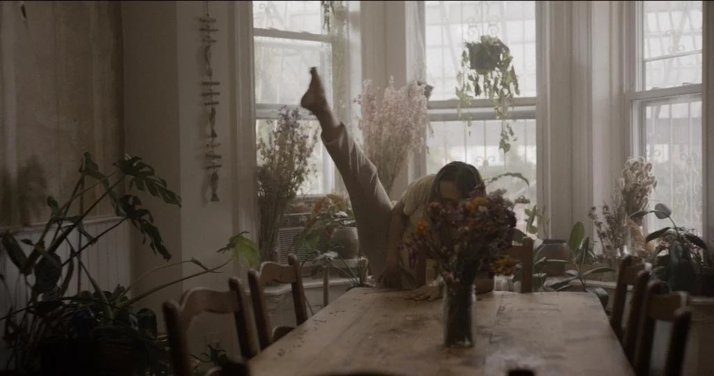 Person stretching on a large wooden dining table surrounded by potted plants, in a bright room with large windows and sheer curtains.