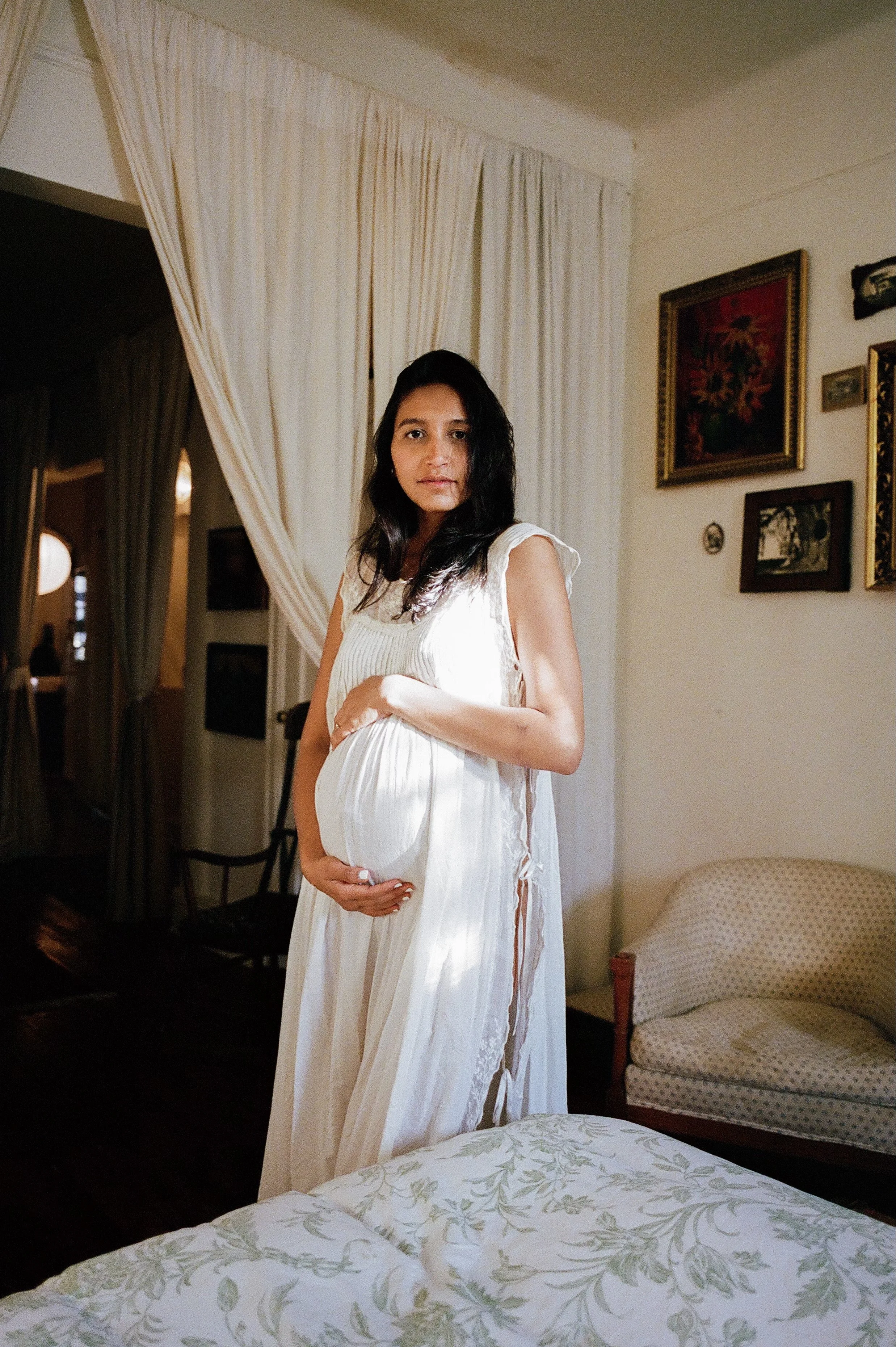 A pregnant woman in a white dress standing indoors, holding her belly with both hands, looking at the camera.