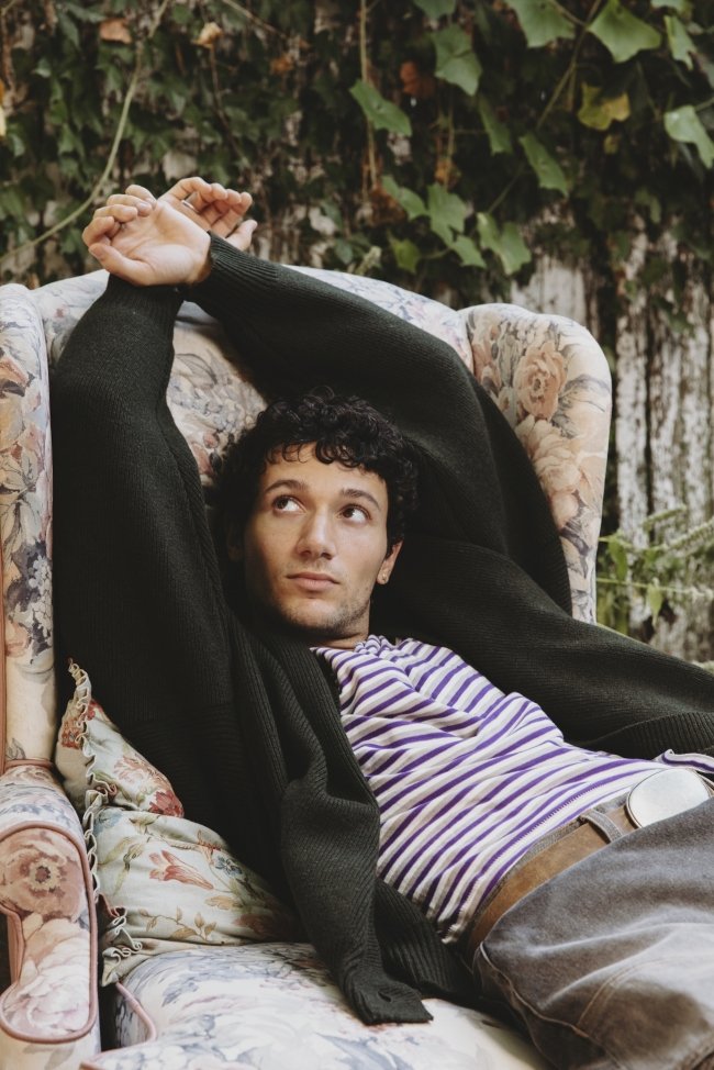 A young man with curly dark hair lying on a floral upholstered couch outdoors, wearing a striped shirt and a black cardigan with his arms stretched above his head, looking off to the side.